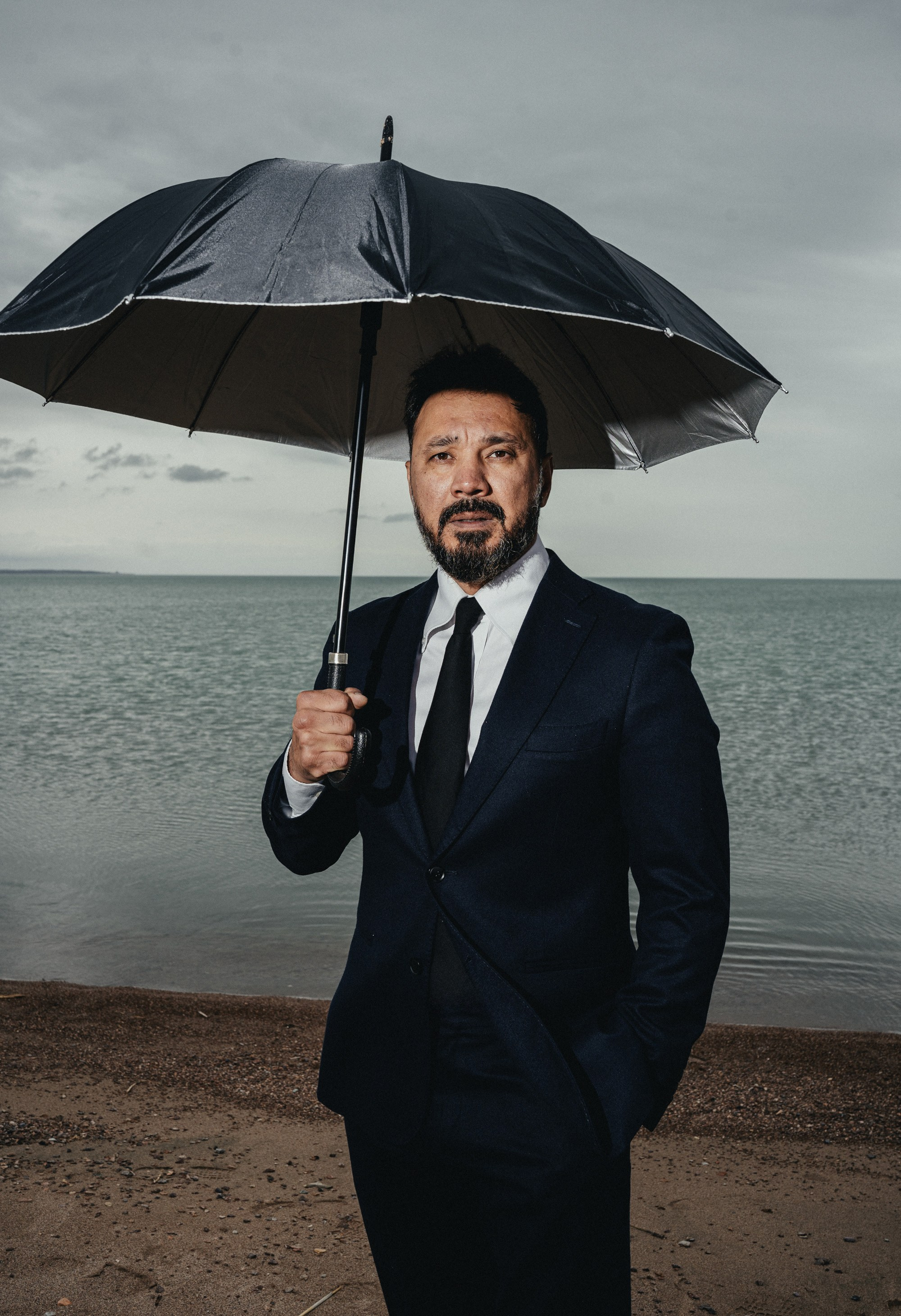 Мужчина в деловом костюме и с чёрным зонтом стоит на берегу моря в пасмурный день