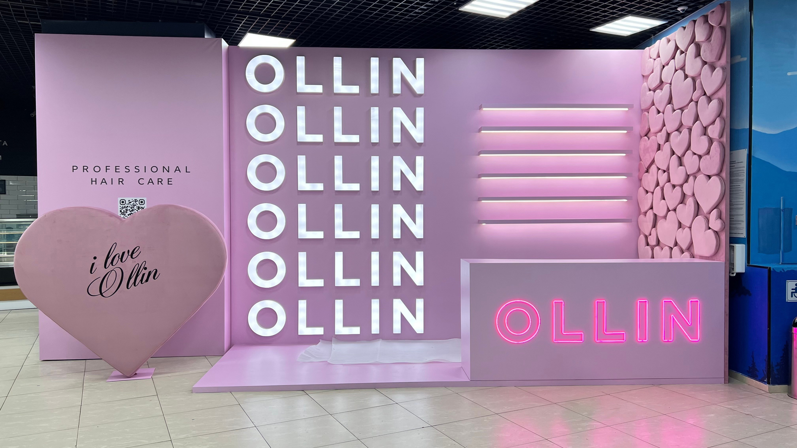 Ярко-розовая минималистичная фотозона бренда «OLLIN PROFESSIONAL HAIR CARE» в интерьере. Центральная стена украшена вертикальной неоновой надписью с шестикратным повторением слова OLLIN. Слева расположено объемное розовое сердце с надписью «i love Ollin», справа — стойка рецепции с яркой розовой подсветкой названия бренда и декоративная панель, полностью покрытая маленькими розовыми сердечками.