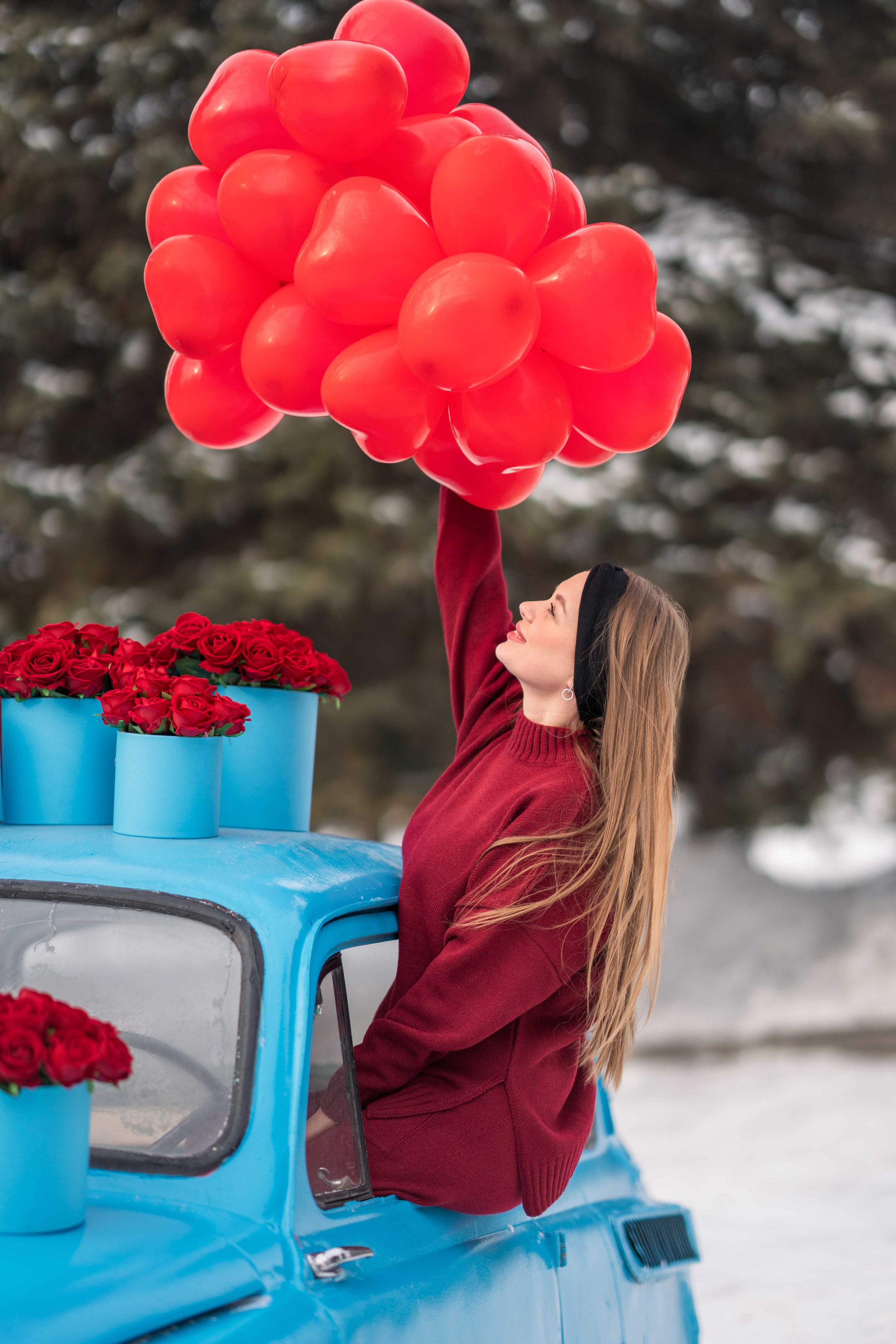 Девушка с красными шариками в виде сердец сидит в окне голубой ретро машинки