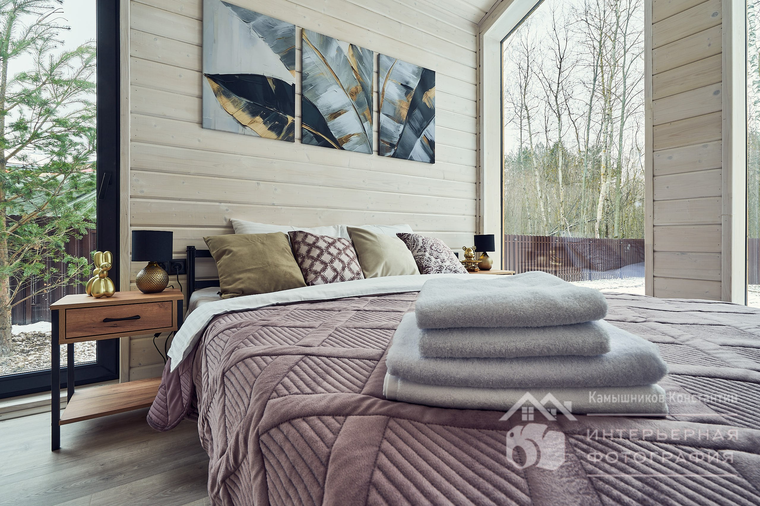 Интерьерная съемка большой спальни в загородном доме в Московской области, где на переднем плане - белые пушистые полотенца, а на заднем - подушки, стена с интерьерной живописью и два панорамных окна, выходящих на разные стороны дома