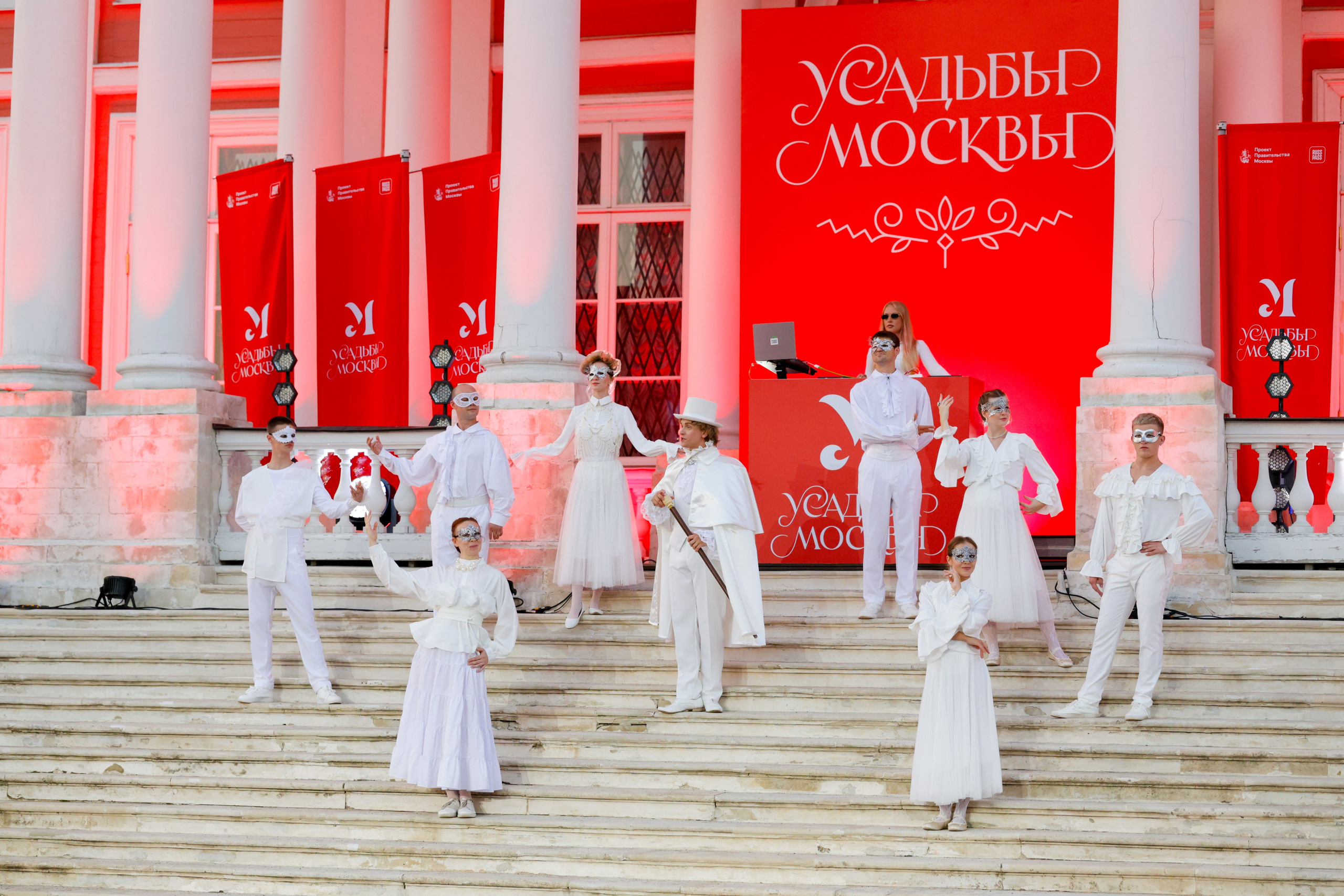 Фотосъёмка концерта и театрального представления на свежем воздухе в парке-усадьбе Кусково для проекта 'Московские усадьбы'. На сцене группа артистов в белых костюмах с масками, стоящих на ступенях перед красным баннером с логотипом проекта. Освещённые светом прожекторов, они исполняют театральное представление. Атмосфера на сцене — торжественная и загадочная