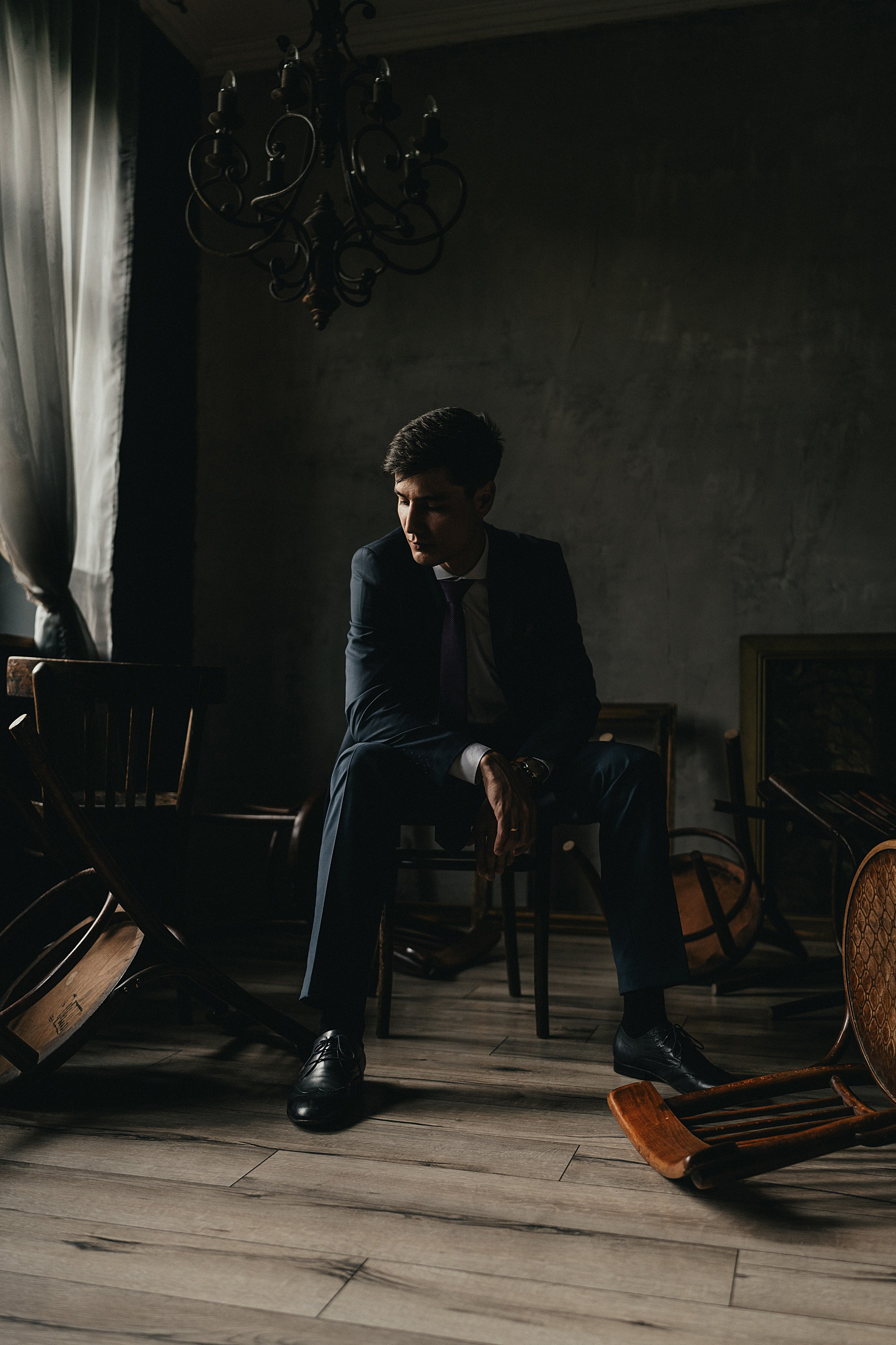 Мужчина в темном костюме сидит среди перевернутых стульев в полуосвещенной комнате