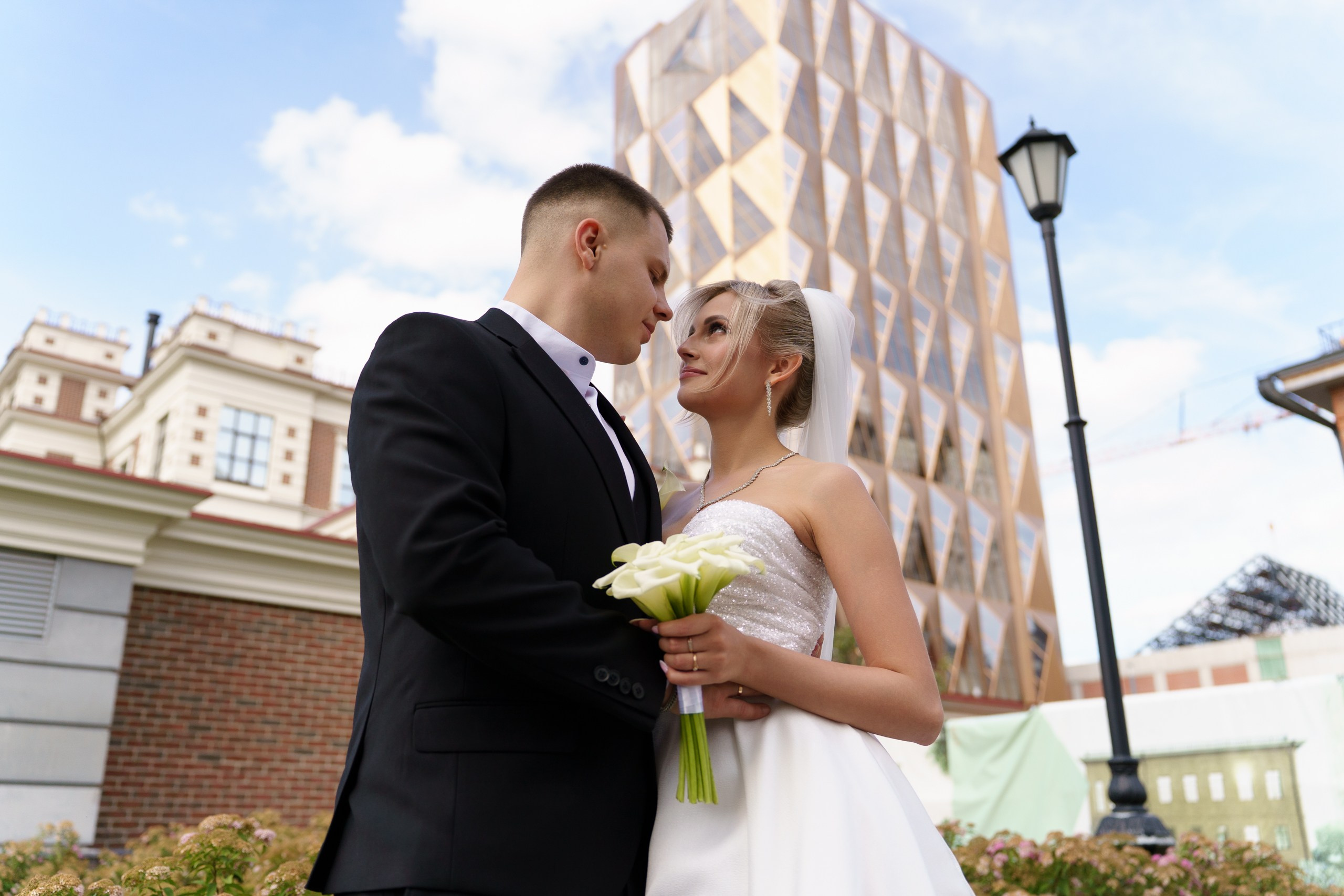 Невеста в белом платье с фатой и жених в чёрном костюме позируют на фоне городского пейзажа. Невеста держит букет белых калл, создавая атмосферу романтики и торжества | профессиональная съемка свадебного торжества Екатеринбург