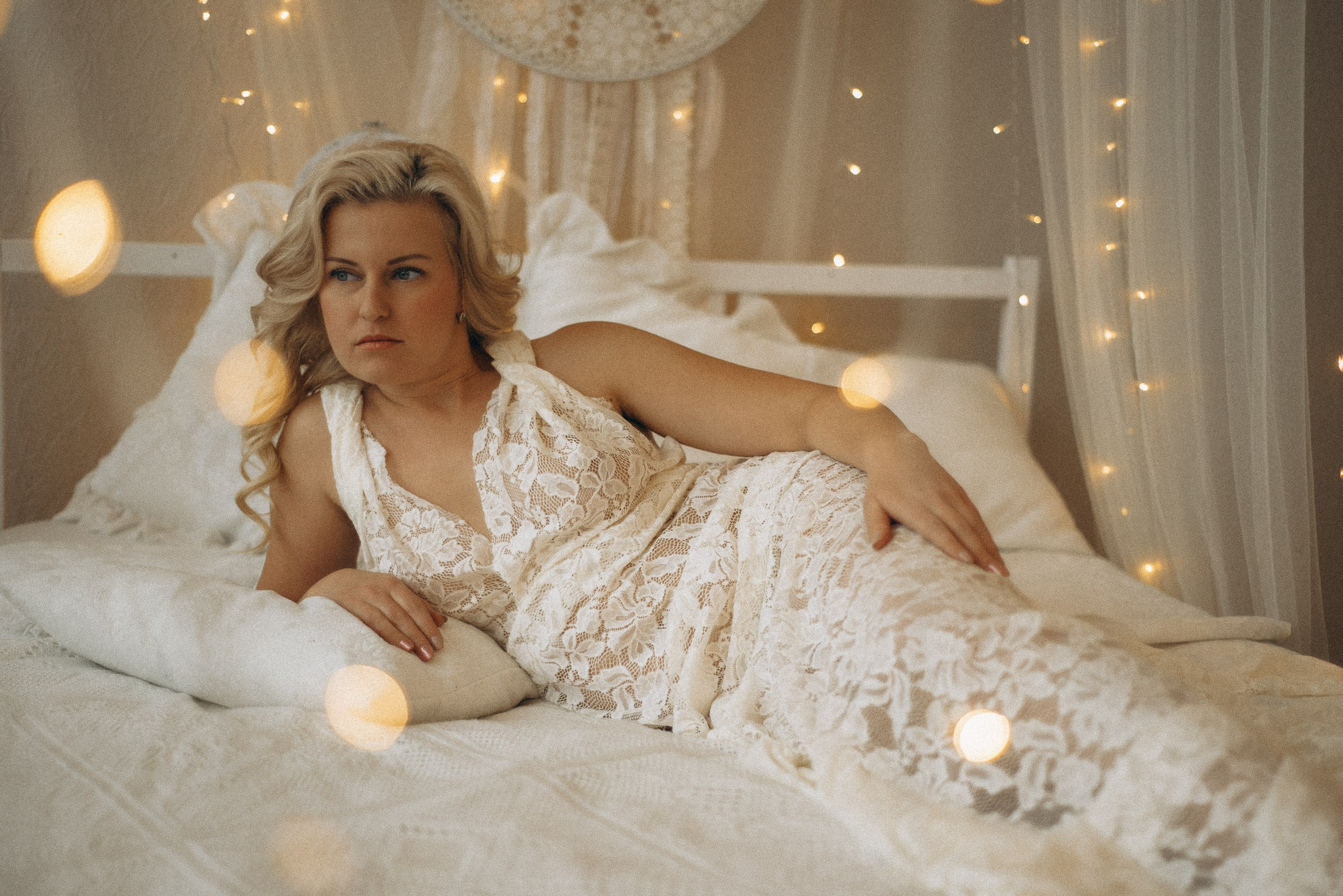 Натали Каролин лежит на боку на кровати в бохо-интерьере. Задумчивый взгляд в сторону, модель в кружевном платье в пол. Теплый свет гирлянд, ловец снов на фоне. Фотограф Василий Дан, Новосибирск