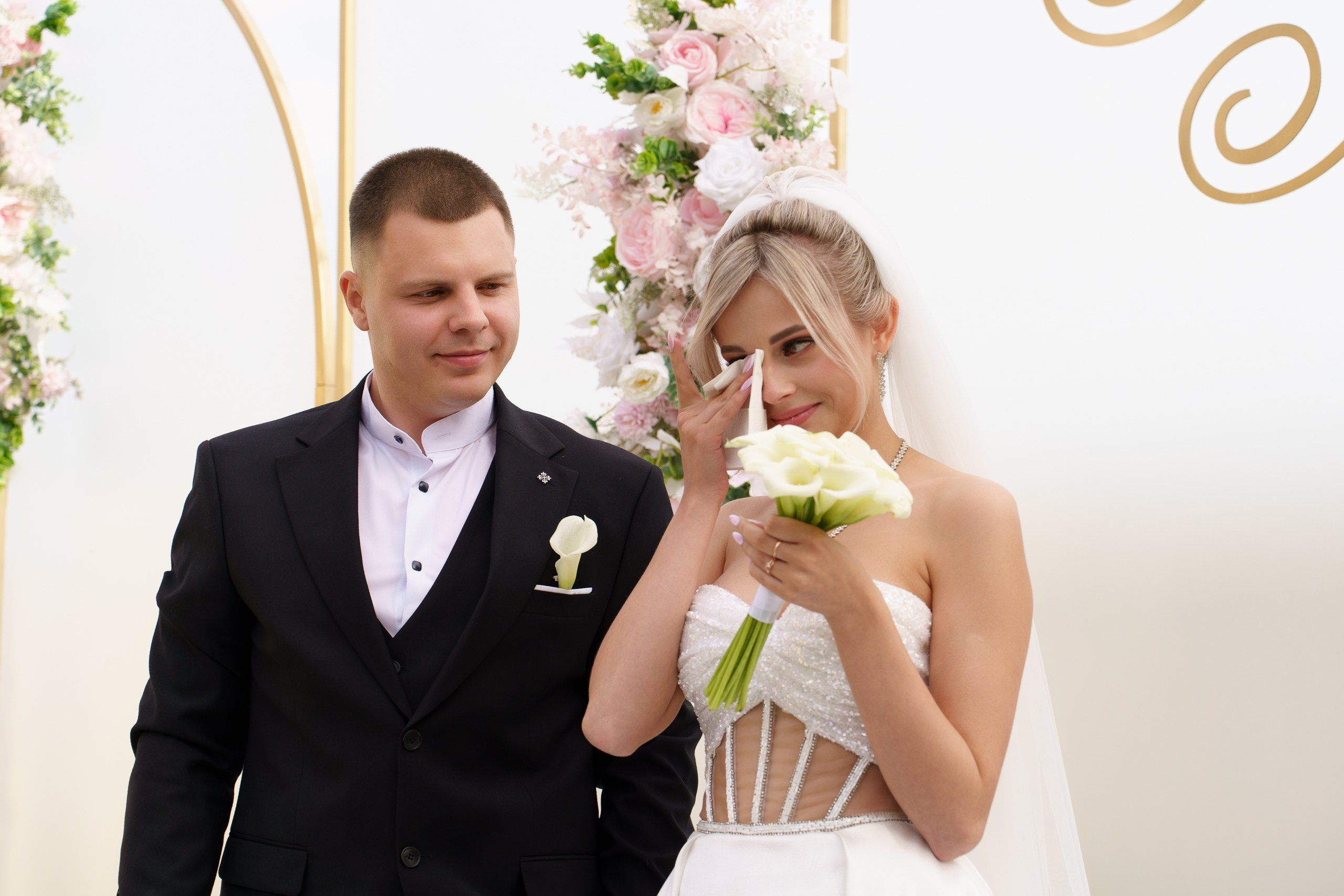 Волшебный момент свадьбы: невеста в роскошном белом платье и жених в стильном чёрном костюме. Трогательная сцена: невеста с букетом калл вытирает слезу счастья, а жених смотрит на неё с нежностью. На фоне — изысканная цветочная композиция | свадебный фотограф Екатеринбург