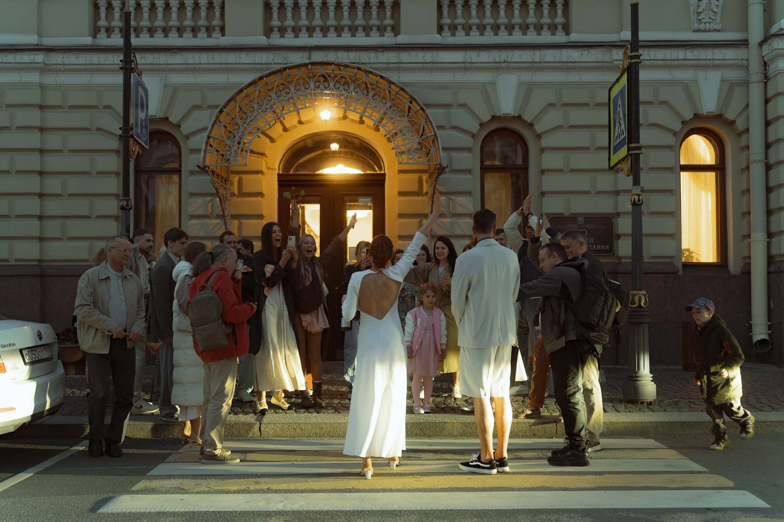 Мария поднимает руку на зебре перед освещённым фасадом ЗАГСа, гости окружают пару