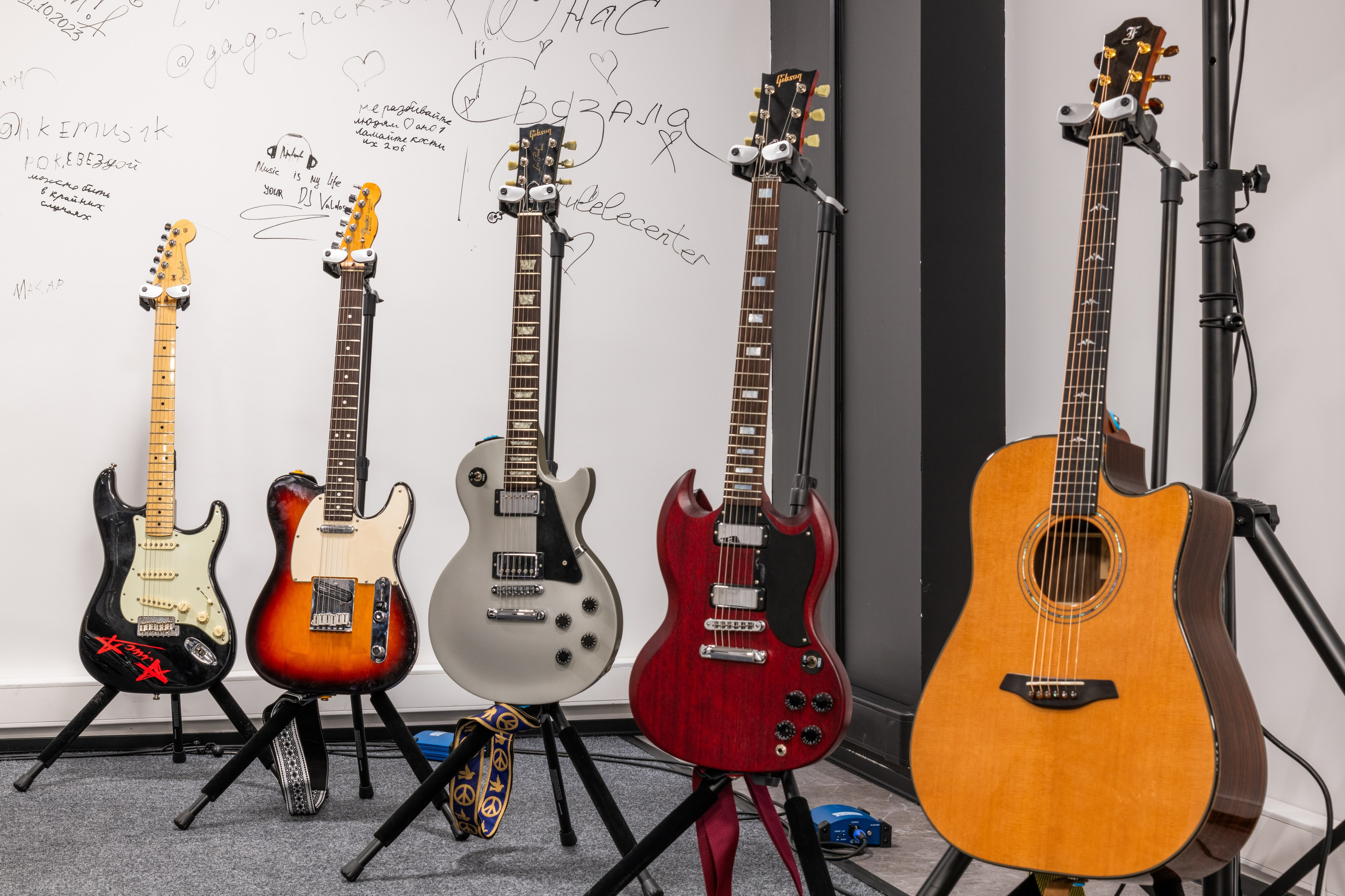 Крупный план электрогитар, выставленных на подставках, с доской для маркеров, покрытой музыкальными нотами и подписями на заднем плане.