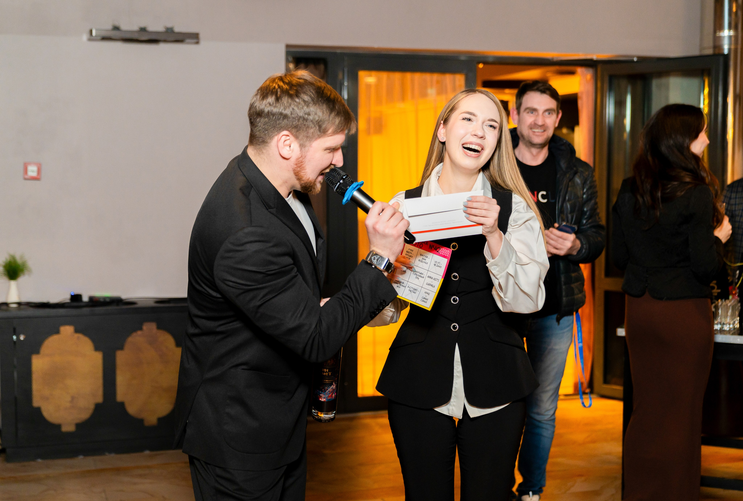 Отель Radisson Sonya. Ведущий в чёрном костюме с микрофоном берёт интервью у смеющейся девушки с конвертом на корпоративном мероприятии. Фотосъёмка событий, фотограф Мальцев Евгений.
