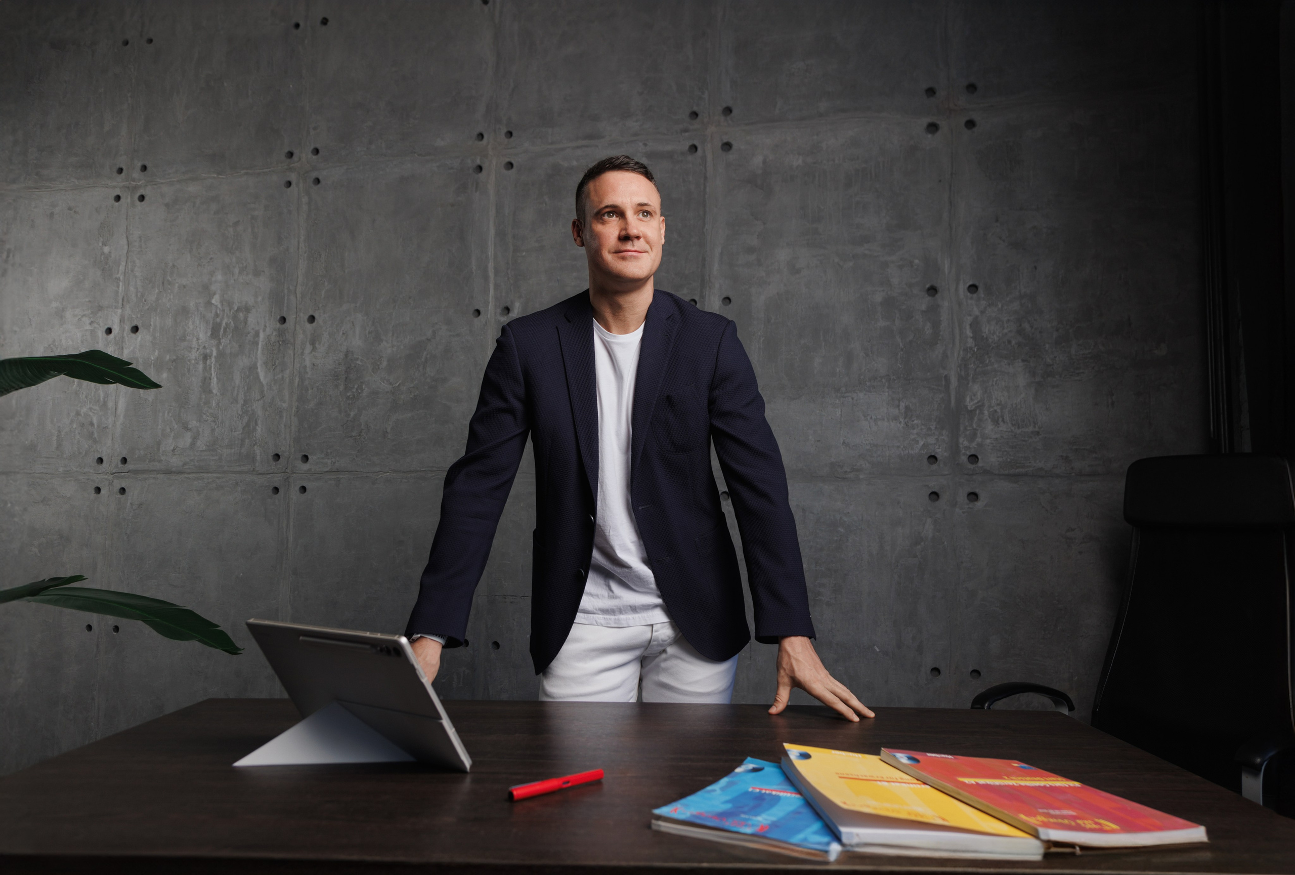 Образ задумчивого педагога: мужчина стоит у стола, опираясь на него, и смотрит в сторону. Естественная поза в студии.