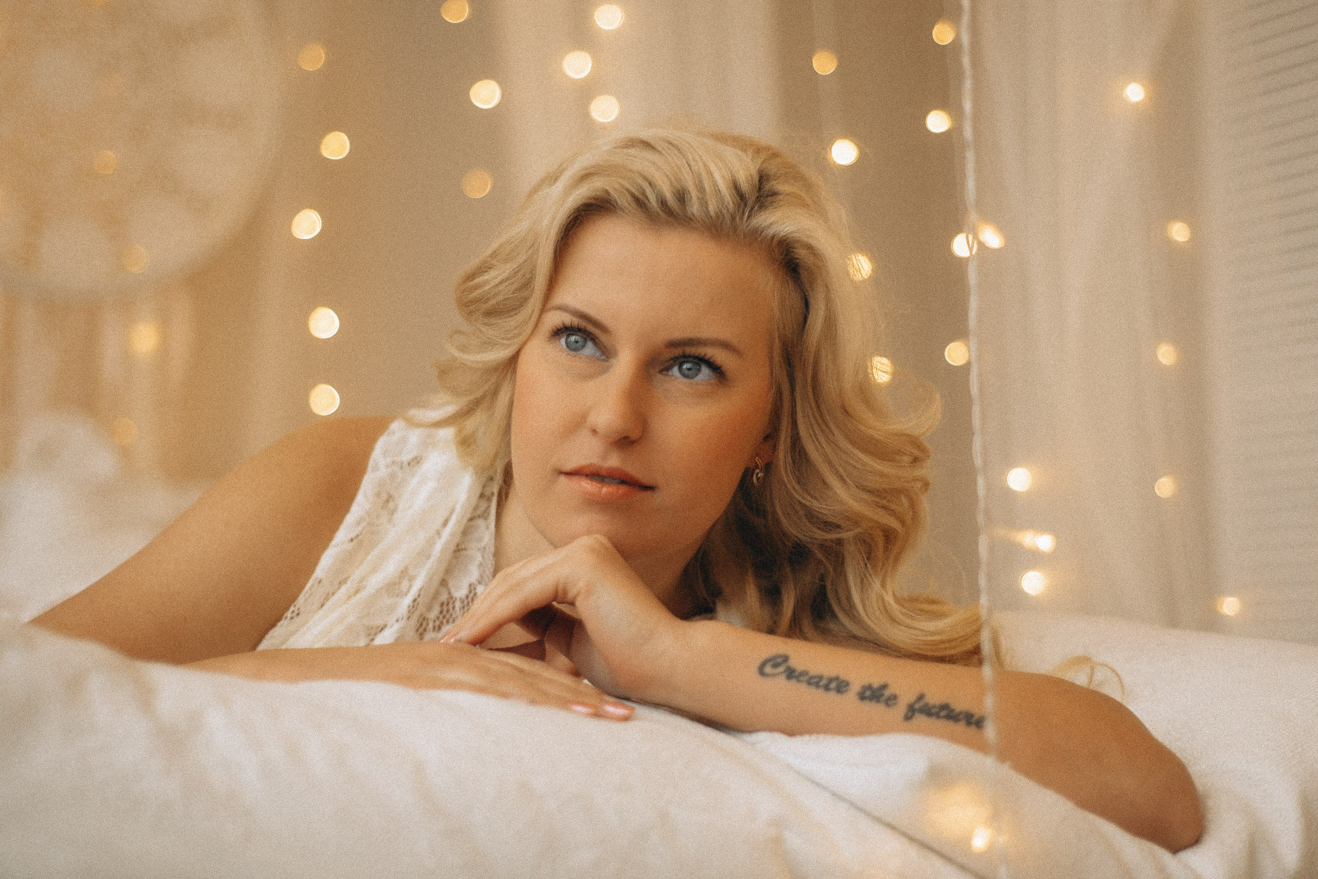 Крупный план Натали Каролин на кровати. Модель в кружевном платье, руки скрещены на подушке, взгляд в кадр. Акцент на голубых глазах и татуировке "Create the future". Фотосессия в стиле бохо. Фотограф Василий Дан