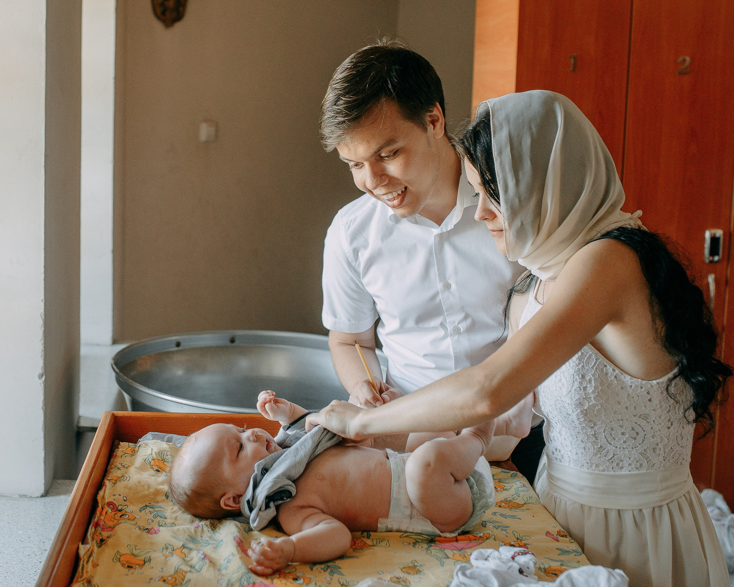 Подготовка к крещению: родители переодевают младенца на пеленальном столике в крестильной комнате.