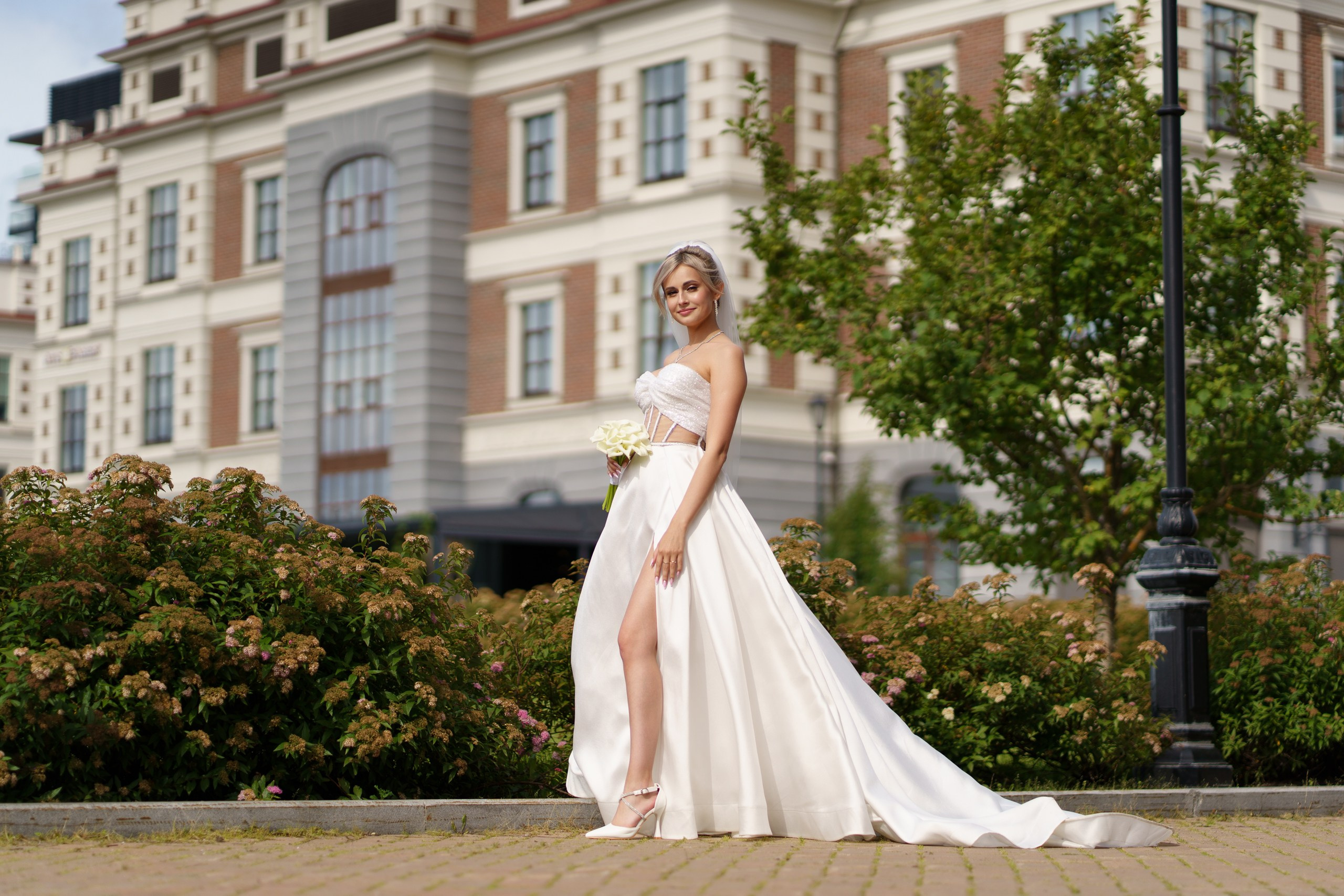 Свадебная фотосессия: невеста в элегантном белом платье с длинным шлейфом стоит на улице. Она держит букет белых цветов, а на заднем плане видны зелёные кусты и здание | фотограф для частных свадеб Екатеринбург