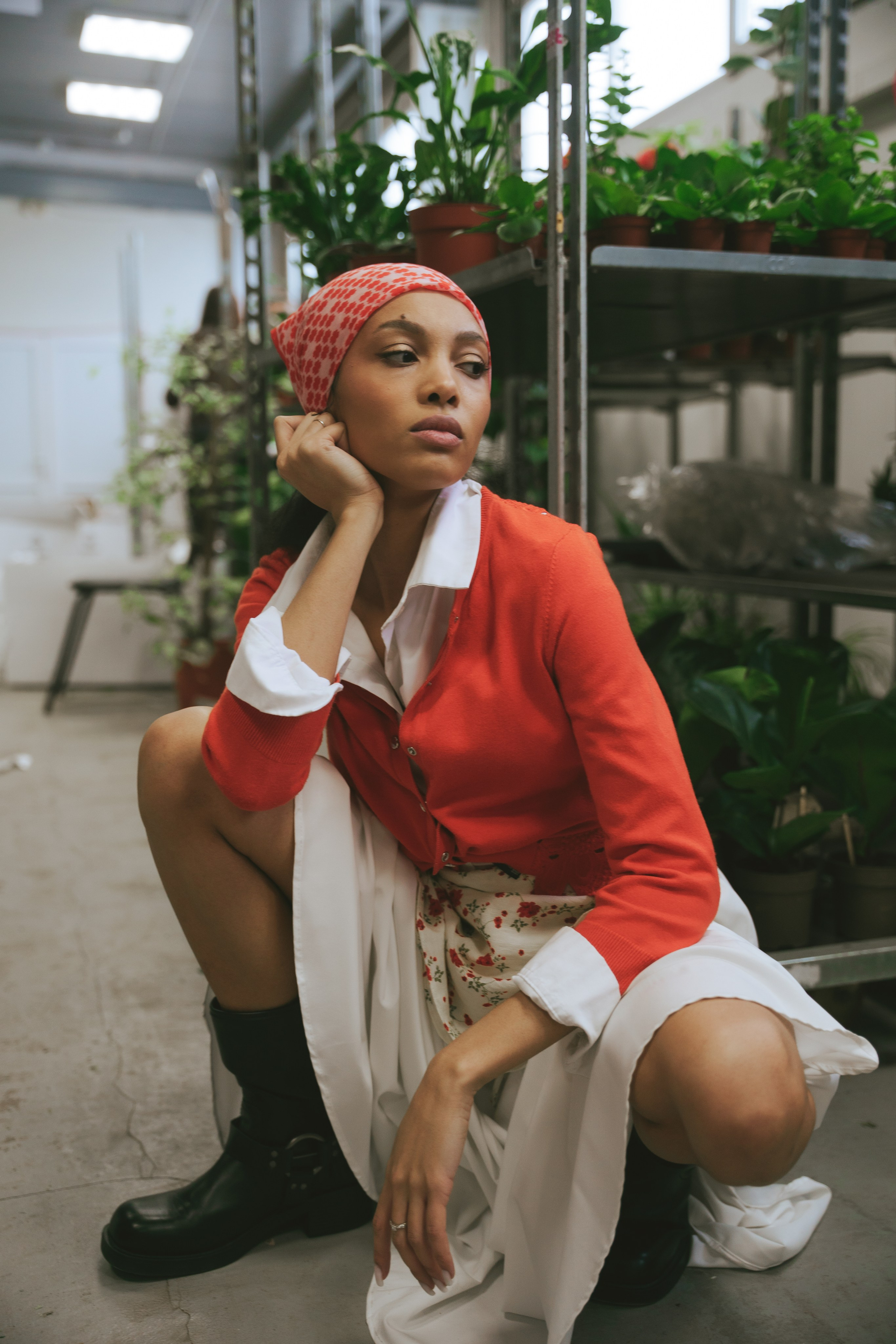 Девушка в ботинках и легком платье сидит на полу среди цветочных стеллажей