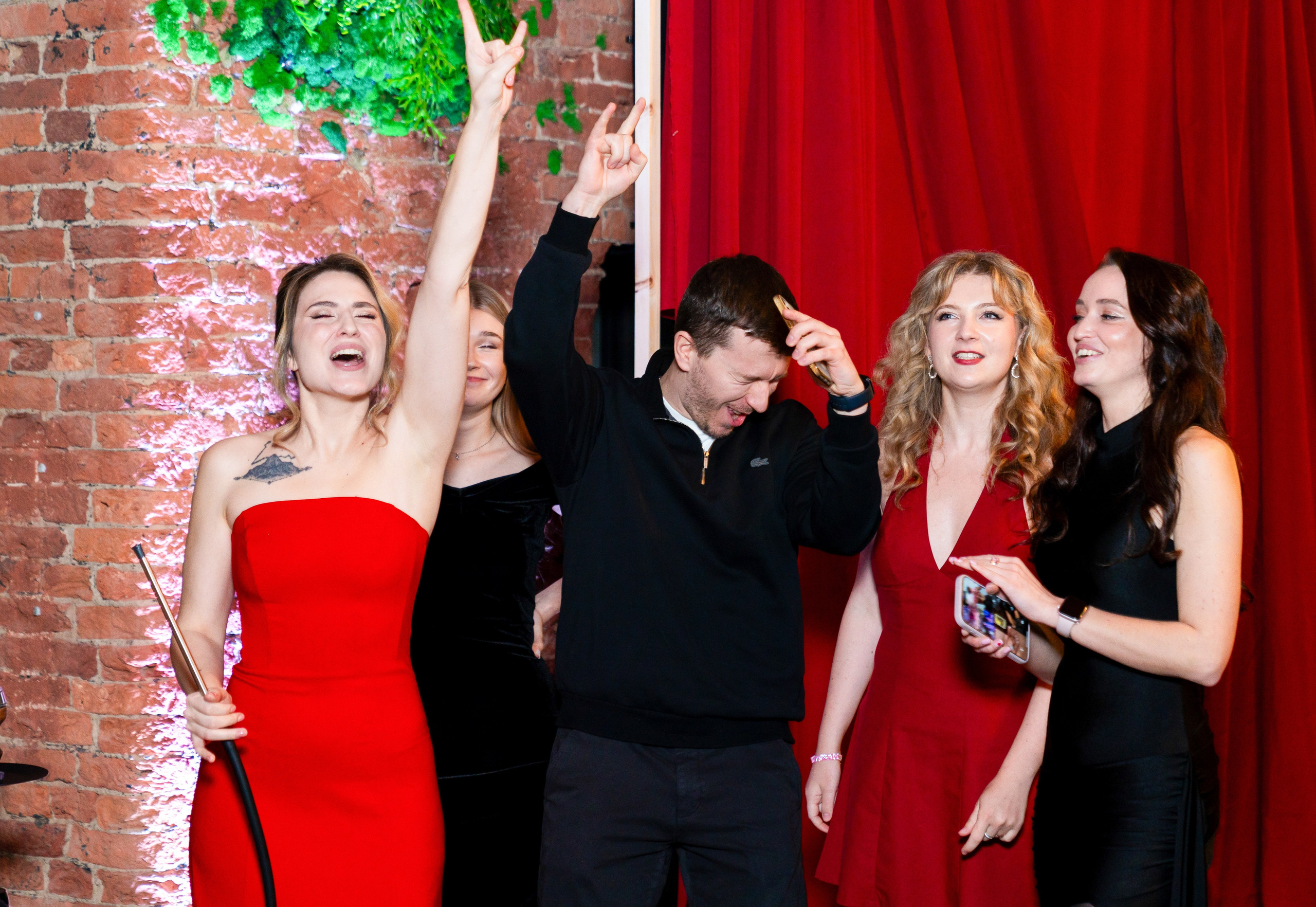 Арсенал лофт. Юбилей. Весёлая компания на празднике у красной шторы: девушка в красном платье показывает рок-жест, мужчина и две девушки рядом. Репортажная съёмка мероприятий, фотограф Мальцев Евгений.