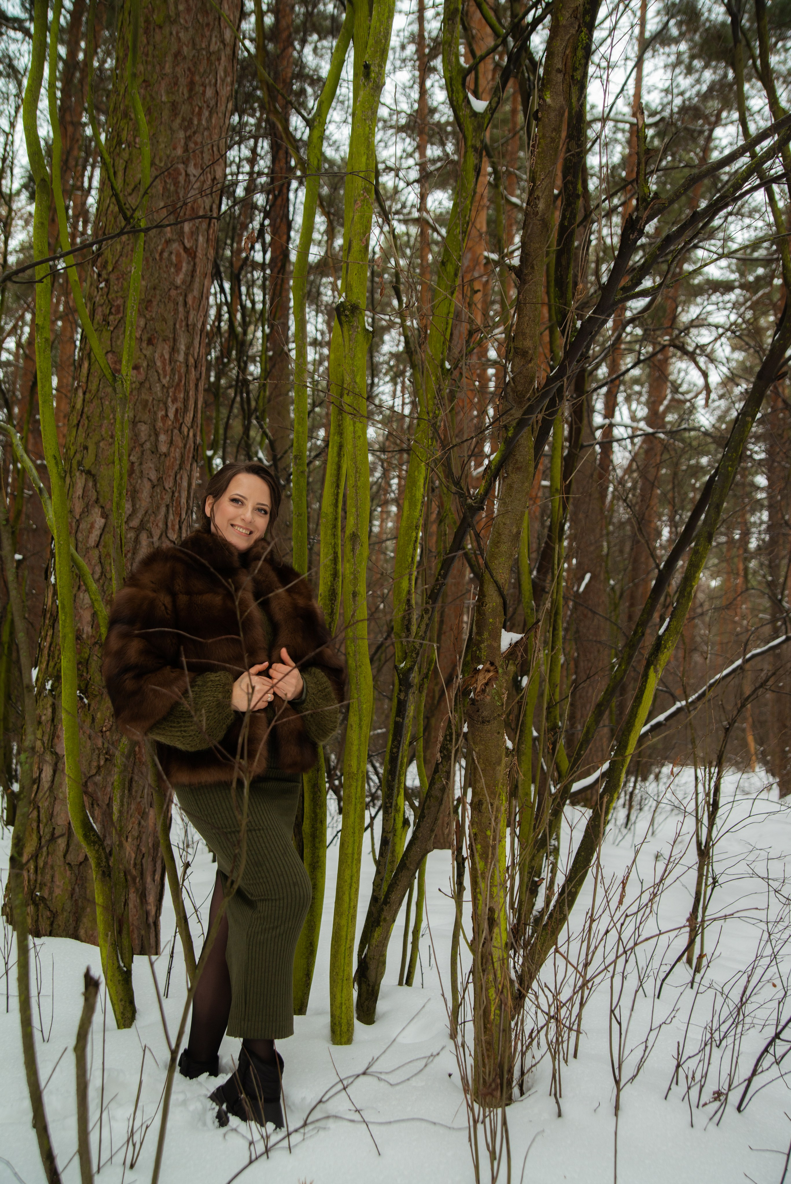 Зимняя фотосессия женщины в зеленом костюме и коричневой шубе в лесу на фоне снега