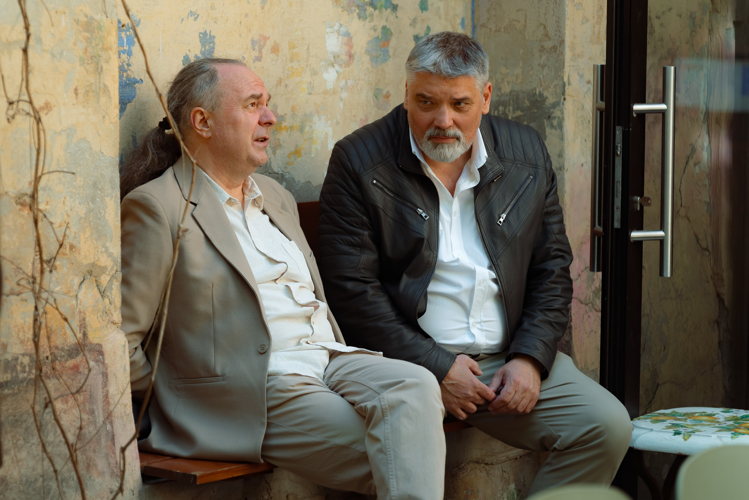 Два гостя в светлых костюмах беседуют на скамейке у каменной стены