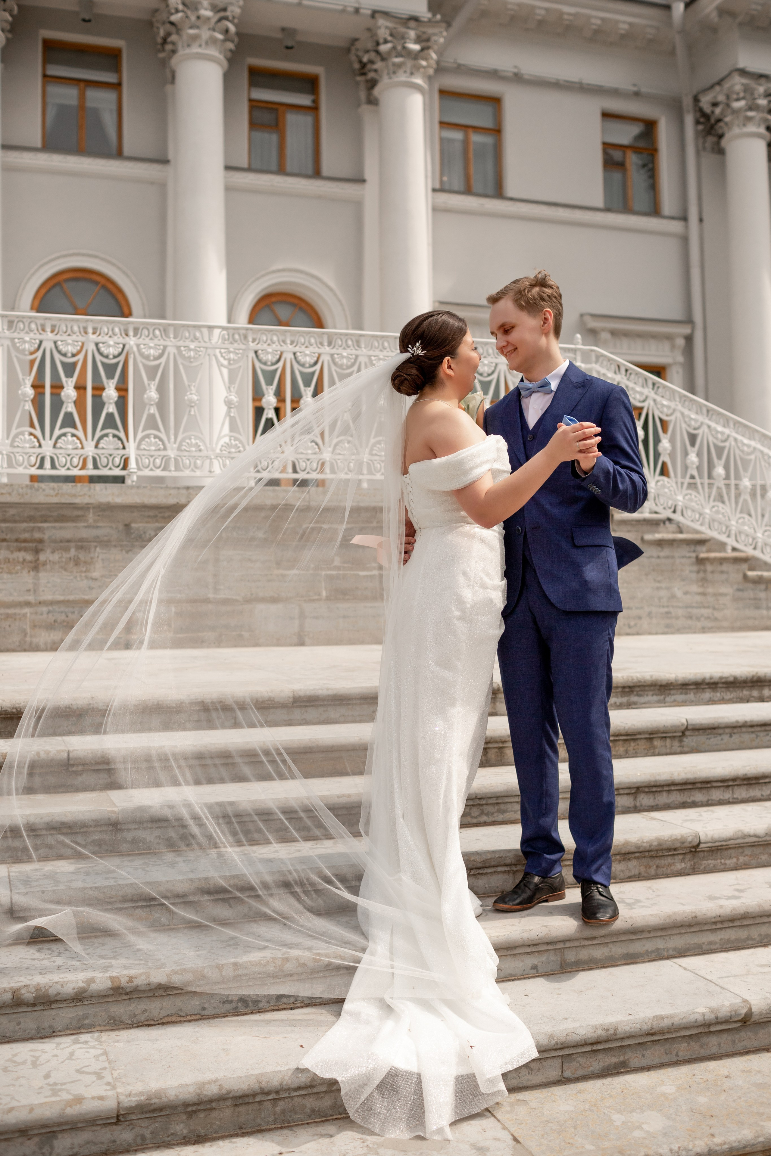 Фата невесты красиво развевается на ветру, когда молодожёны стоят на лестнице около дворца.