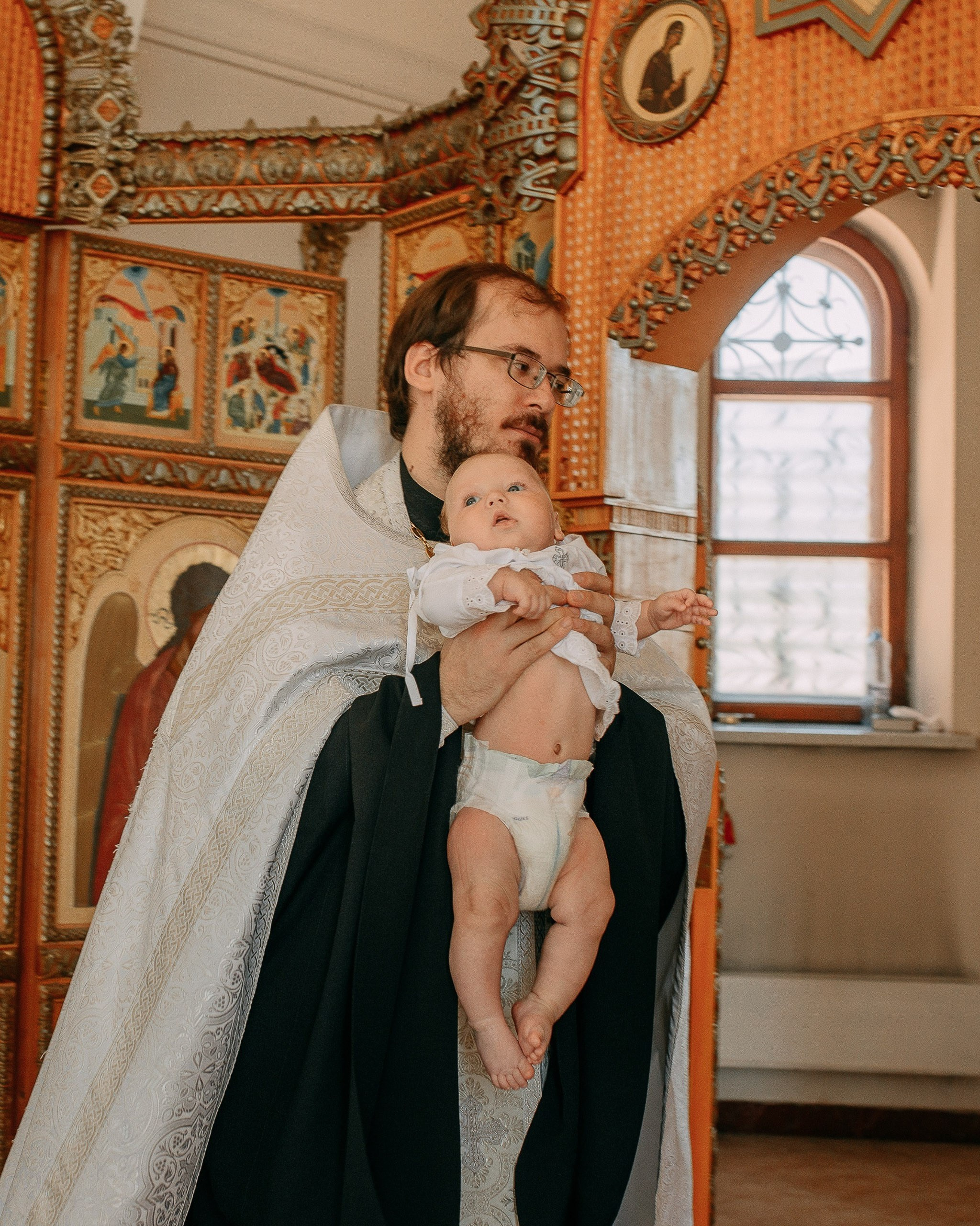 Фотосъемка крещения ребенка, портрет священника с младенцем на руках перед иконостасом. Фотограф на крещение в Новосибирске.
