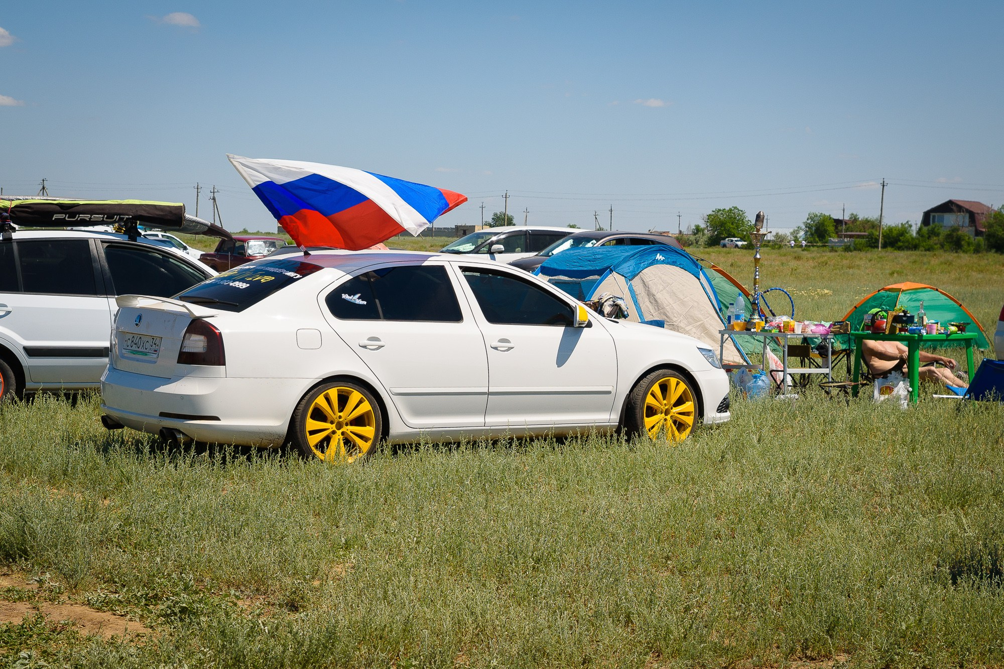Белый автомобиль с жёлтыми дисками стоит на зелёной траве. Сзади автомобиля развевается флаг России