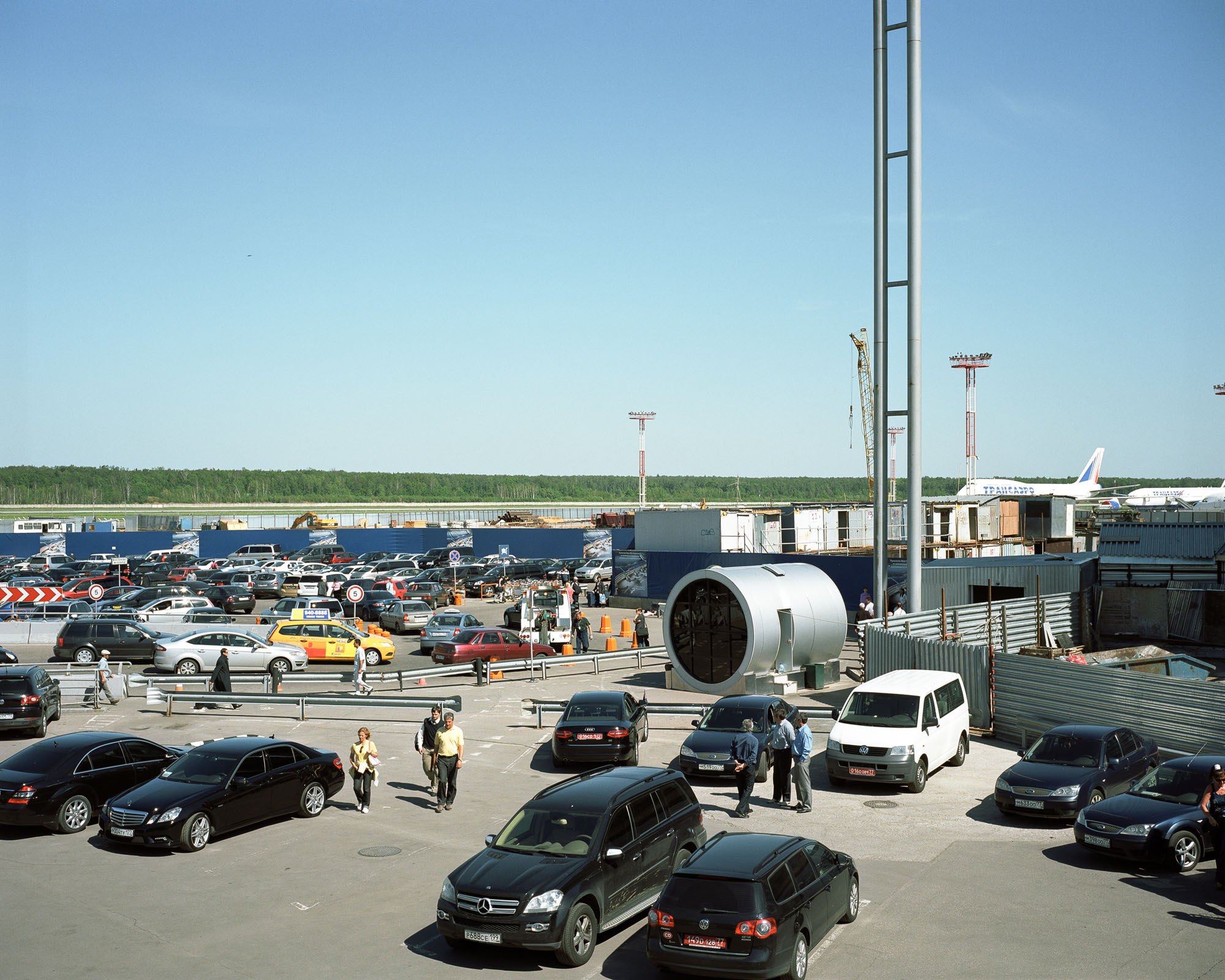 PLATE 25: Parking lot, figures of people, cars, lighting towers, crawler crane, assorted heavy machinery, assorted barriers, corrugated sheet fence, freight containers, kiosk, jet planes