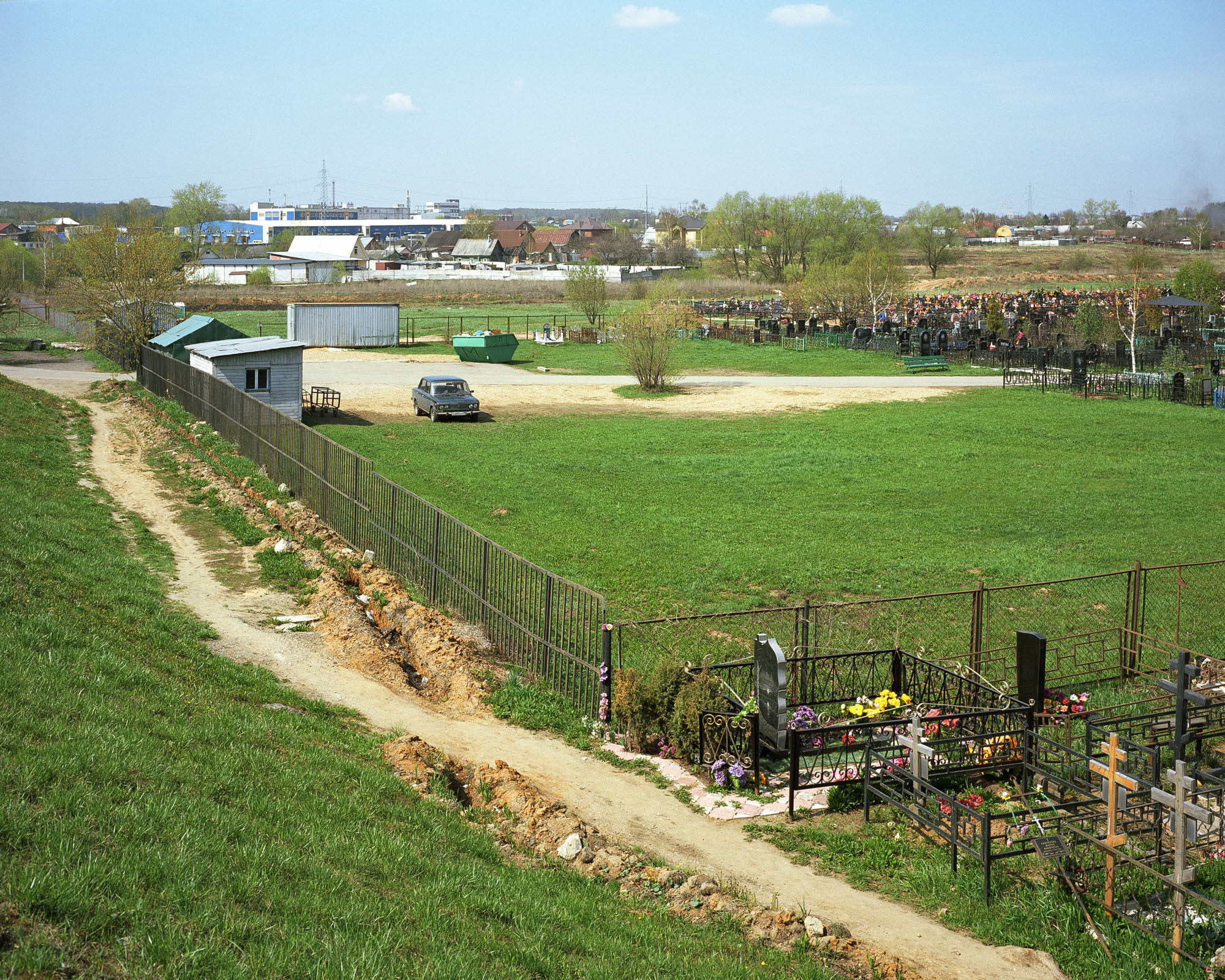 PLATE 19: Grass, assorted barriers, assorted temporary structures, car, trees, graves, paved and unpaved pathways (c/w stones), ditch, waste skip, summer houses