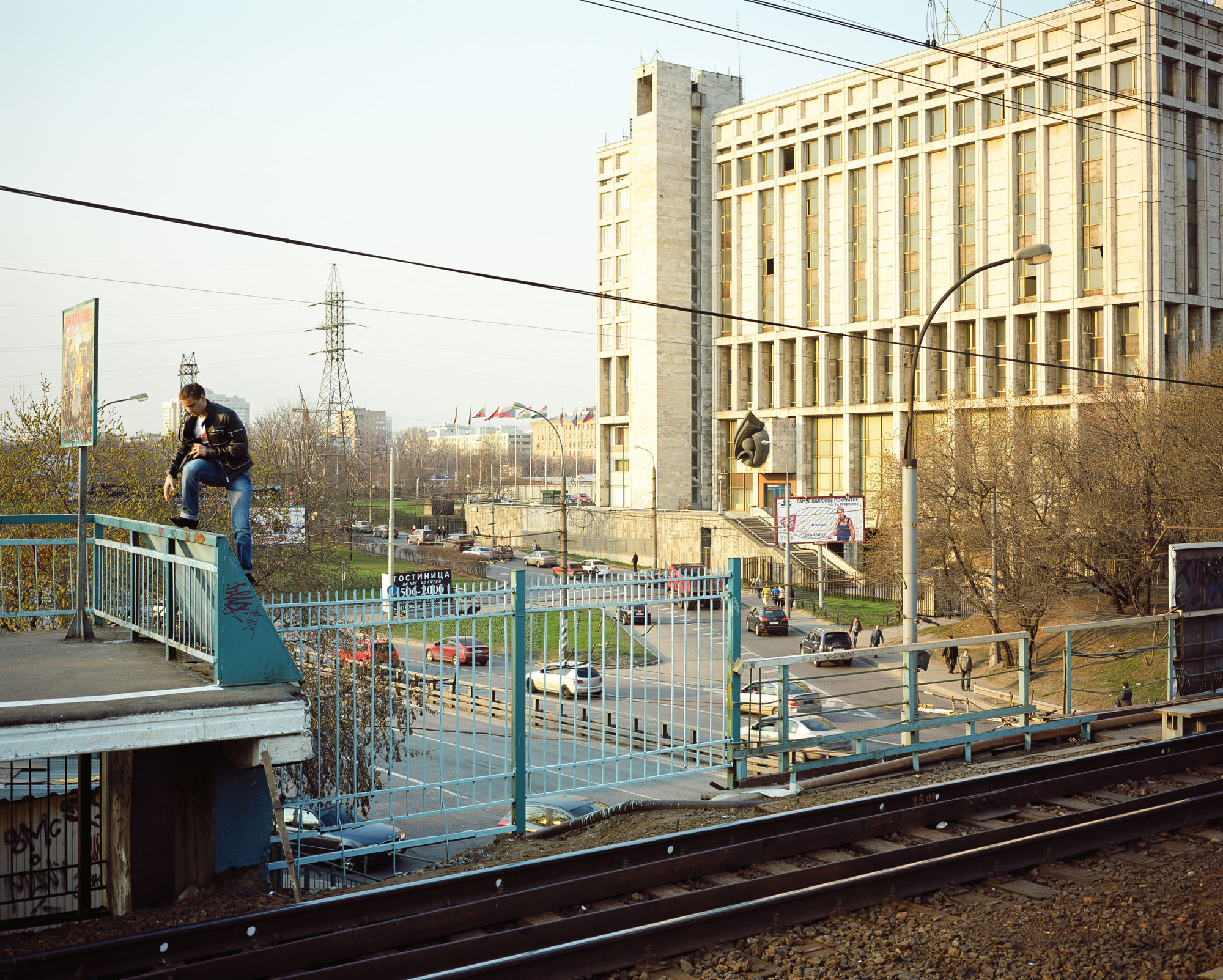 PLATE 5: High voltage tower, non-residential building, assorted barriers, railway platform, railway tracks, crushed rock, lamp posts, flagpoles c/w flags, power lines, figures of people, cars, trees, billboards