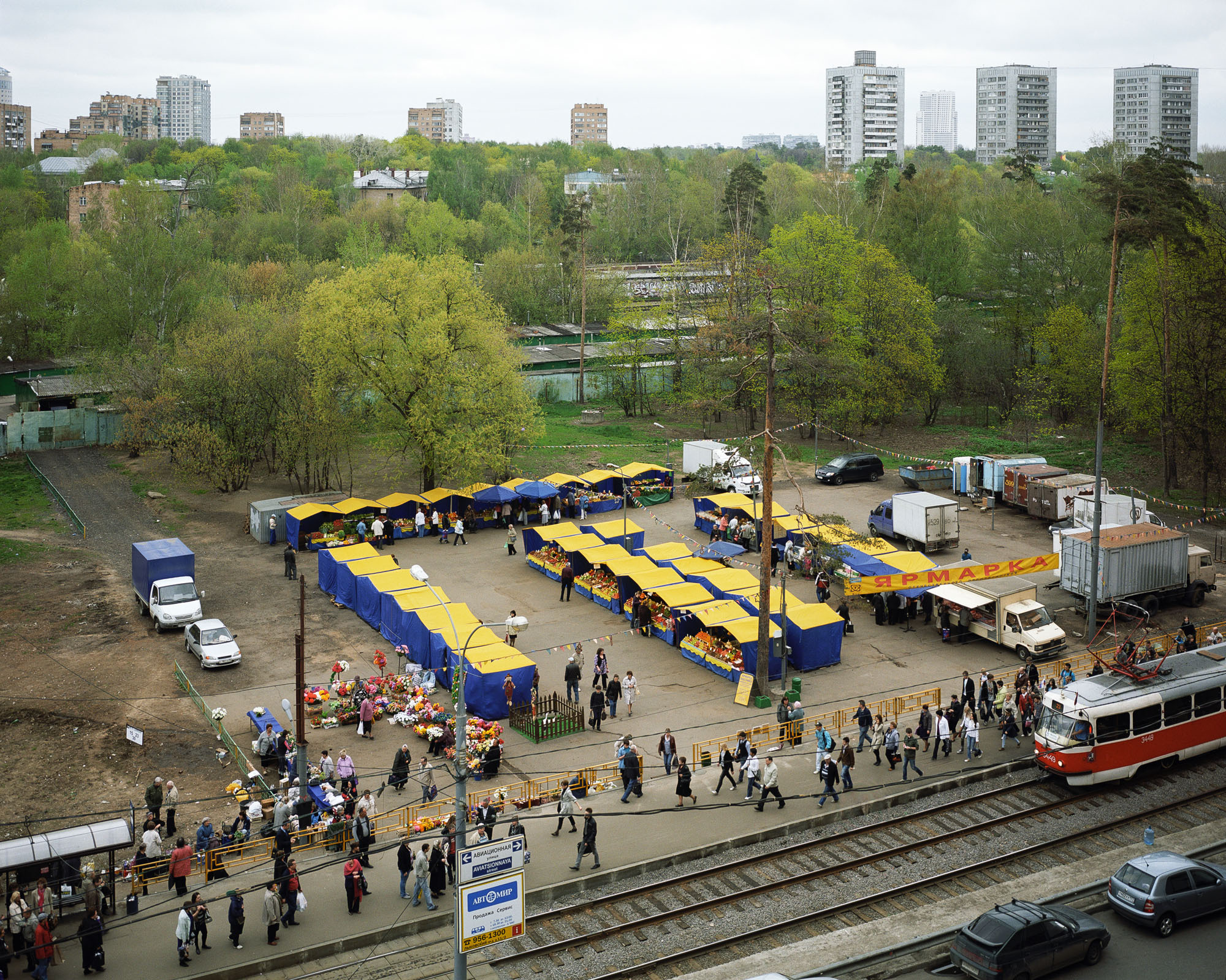 PLATE 43: Market tents, trucks, tram stop c/w shelter and barriers, tram tracks with tram car, figures of people, waste bins (flower pots), tables and display cases with produce, flag decorations, mobile kiosk, banner, trees, car shelters, lamp posts, power lines, residential buildings, figures of people, cars
