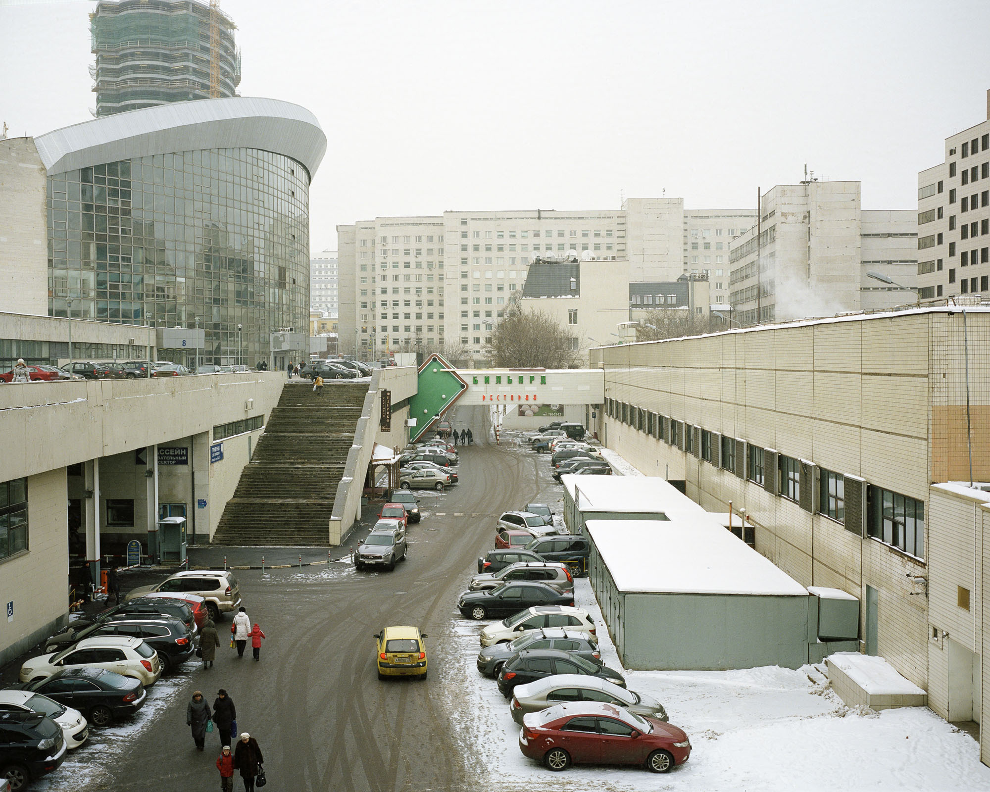 PLATE 20: Sports venue with glass façade, office and utility buildings, metal car shelters, speed bump, barrier posts, stairs, snow patches, figures of people, cars, surveillance cameras, assorted signs