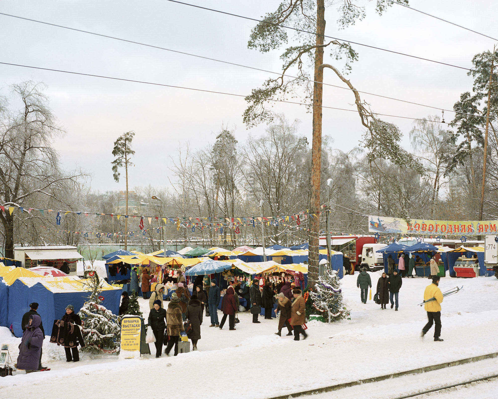 PLATE 39: Market tents, mobile kiosks, tram tracks, figures of people, waste bin, flag decorations, banner, Christmas tree, lamp post, tables and display cases with produce, trees, power lines, snow