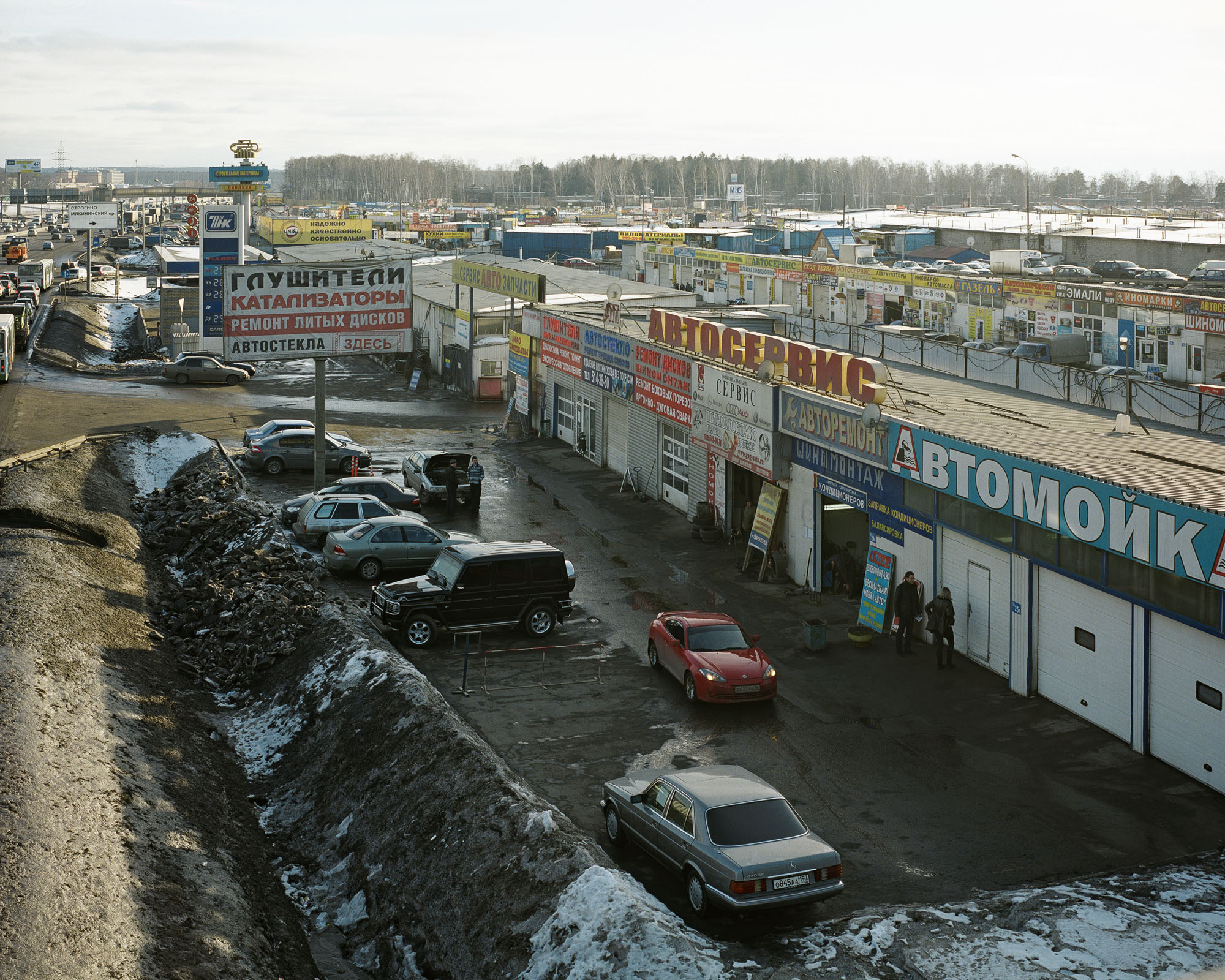 PLATE 27: Parking space c/w assorted cars, figures of people, semi-permanent shop buildings (with A/C units, signage, satellite dishes), filling station, pedestrian bridge, billboards and banners, multi-lane freeway c/w drainage channel, trees, barriers