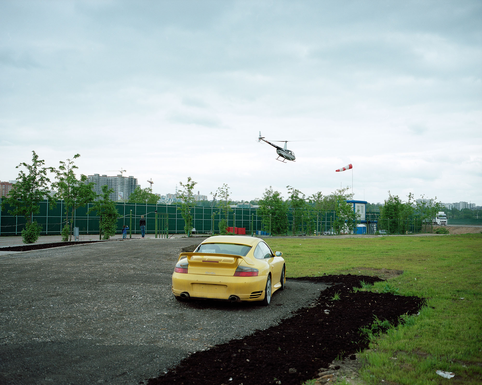 PLATE 24: Нelipad c/w fence, windsock, rubbish bins, portable offices and toilet, helicopter during take off, trees, parking space c/w sports car, figures of people, lawn, truck