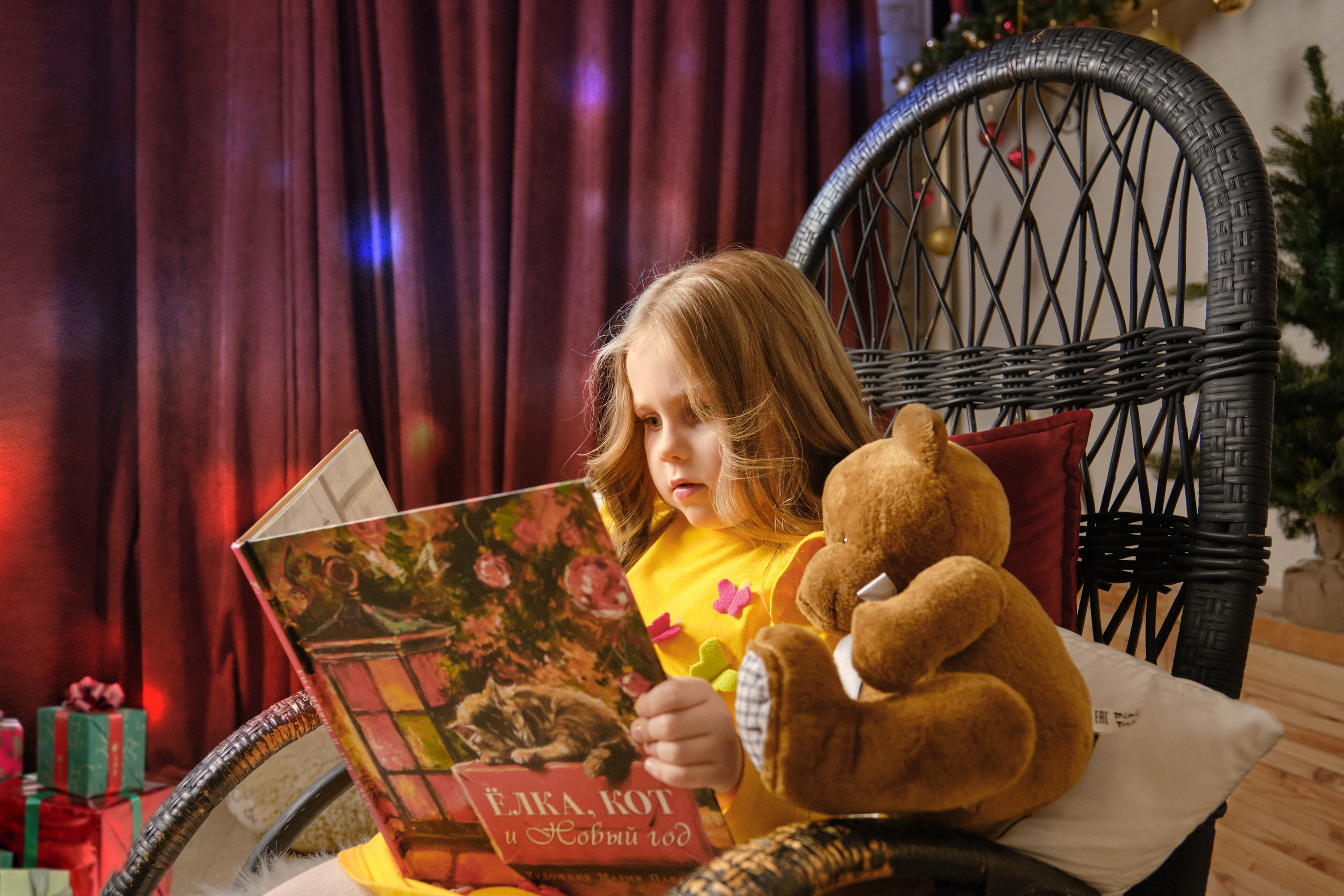 детская новогодняя фотосессия в студии - девочка в новогоднем интерьере сидит в кресле качалке и читает книжку мишке