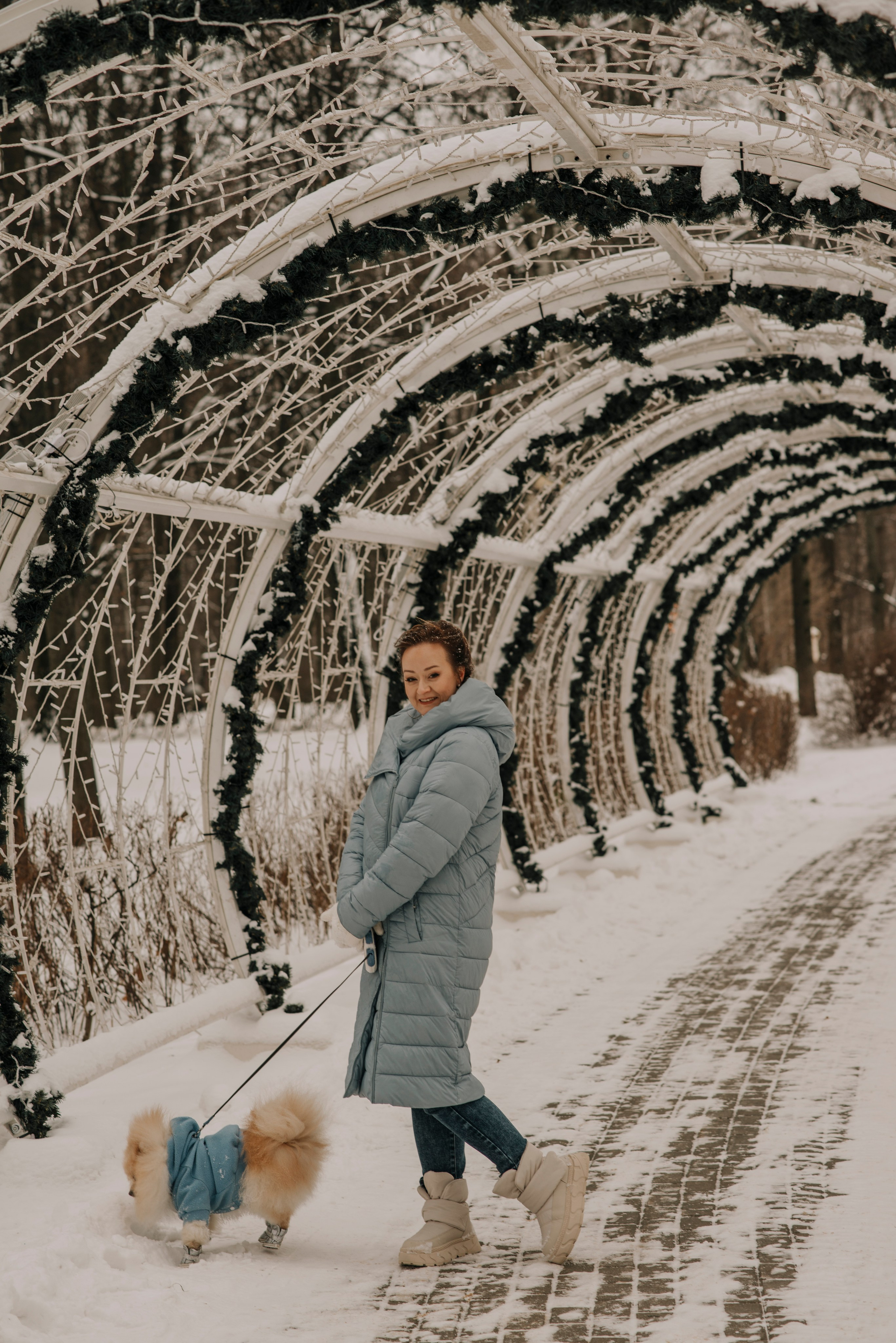 Фотосессия с собакой зимой в заснеженном парке. Рыжеволосая девушка в голубом пуховике держит на поводке шпица