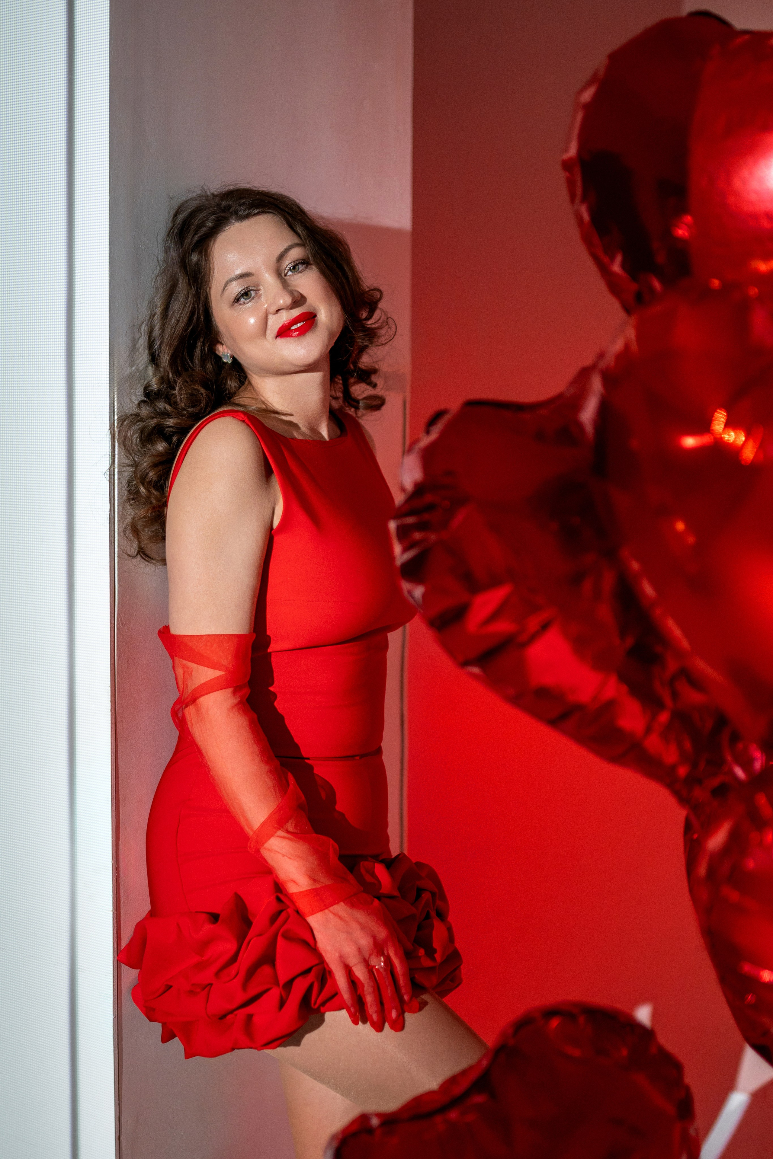 Девушка в красном платье у белой стены с красными воздушными шарами - сердцами