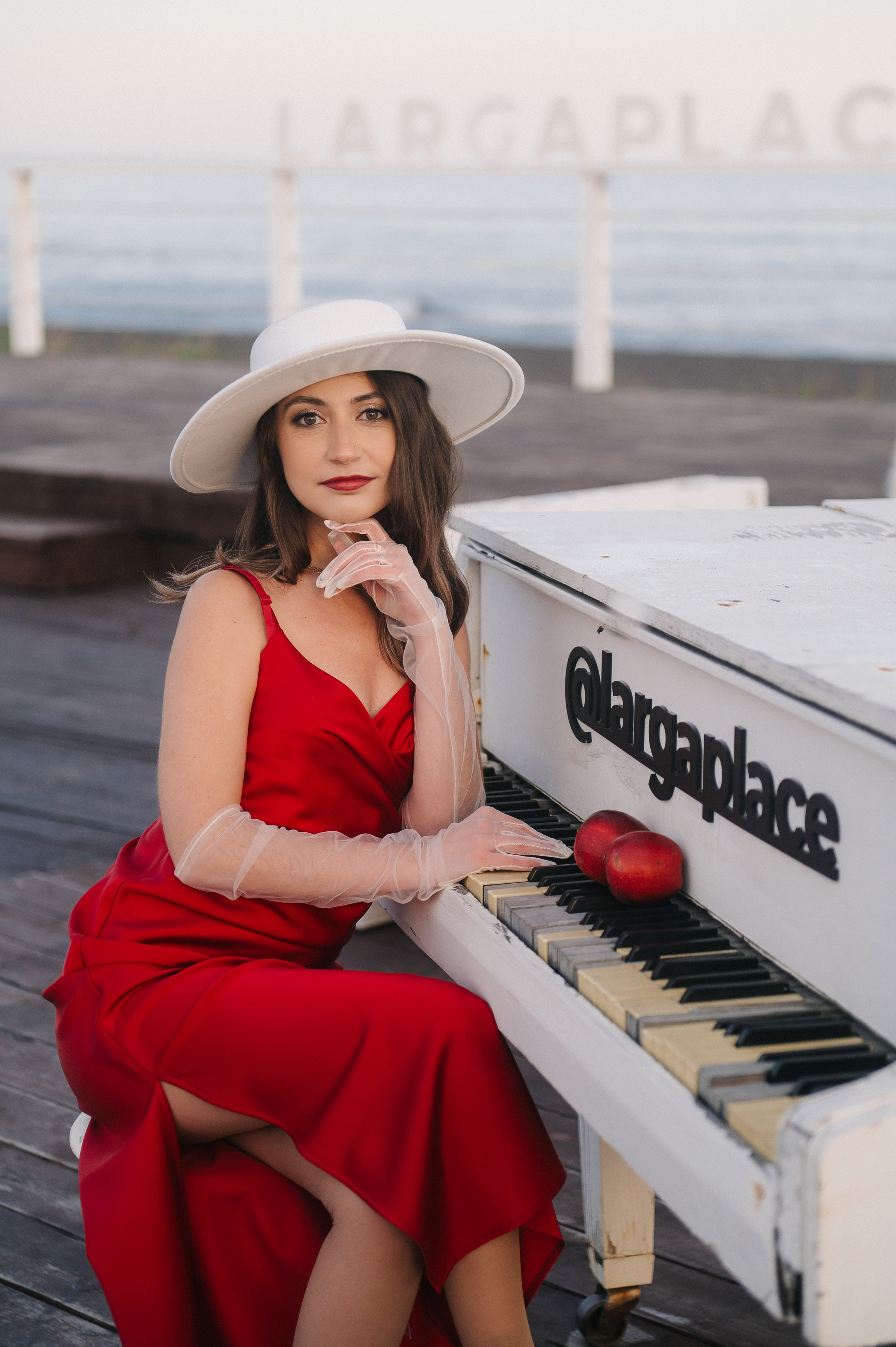 Девушка в красном платье и белой шляпе сидит у белого пианино. На клавишах — два красных яблока. Атмосфера стильная, романтичная, у воды.