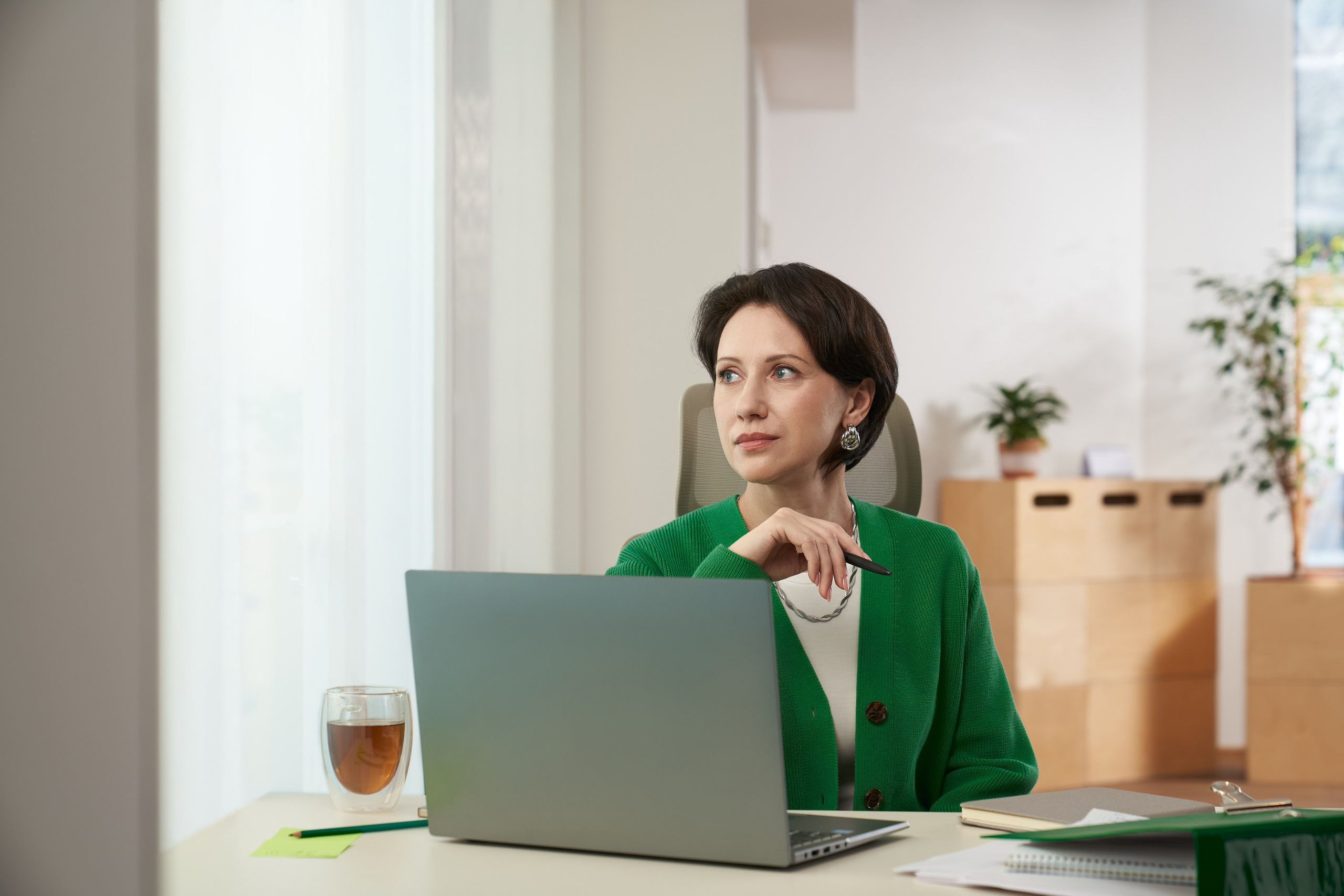 Женщина в зелёном кардигане работает за ноутбуком в офисе, задумчивый взгляд в сторону