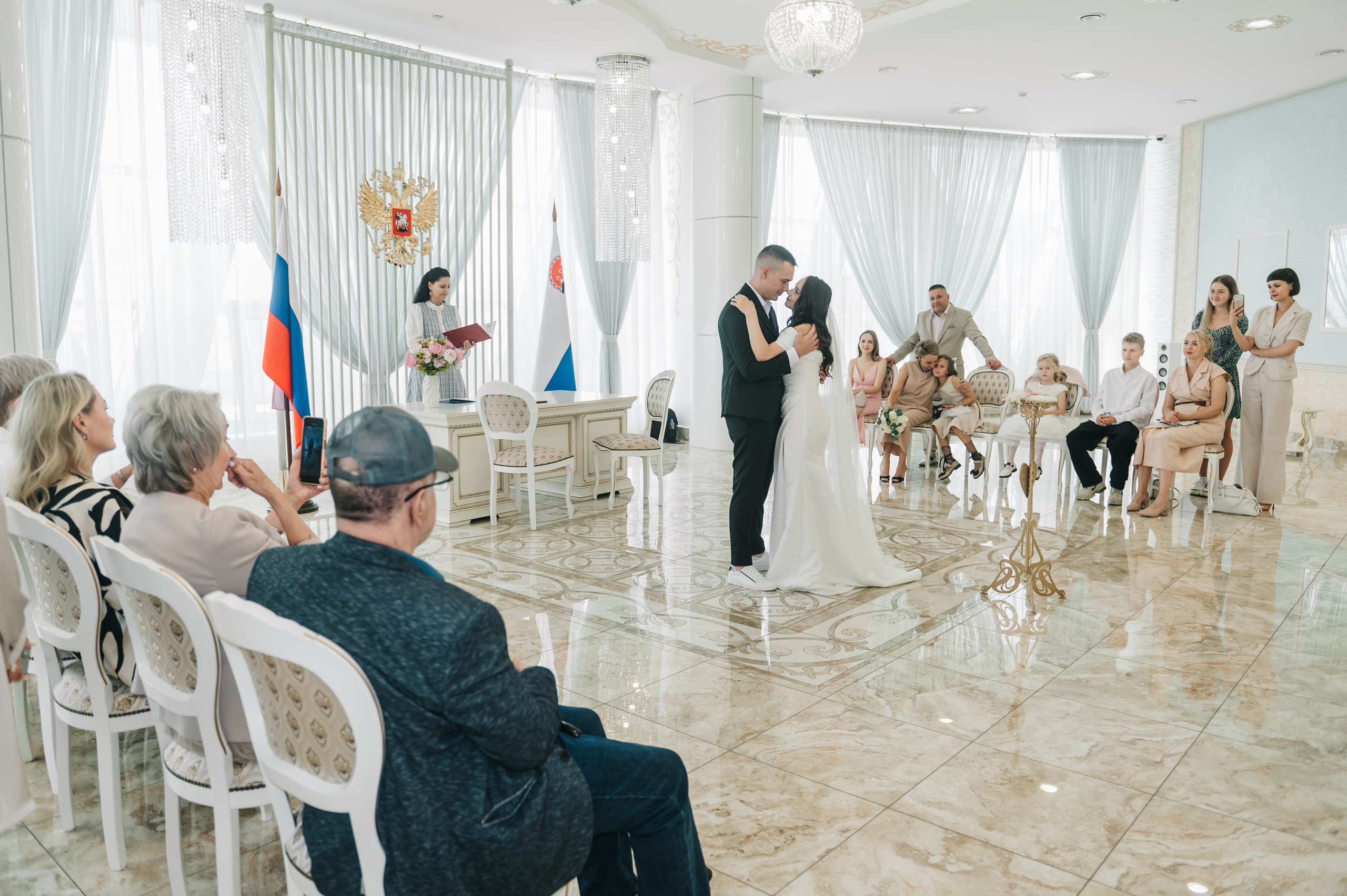 Жених и невеста обнимаются у алтаря в ЗАГСе, гости сидят вокруг. Интерьер светлый, торжественный, флаги и герб на фоне.