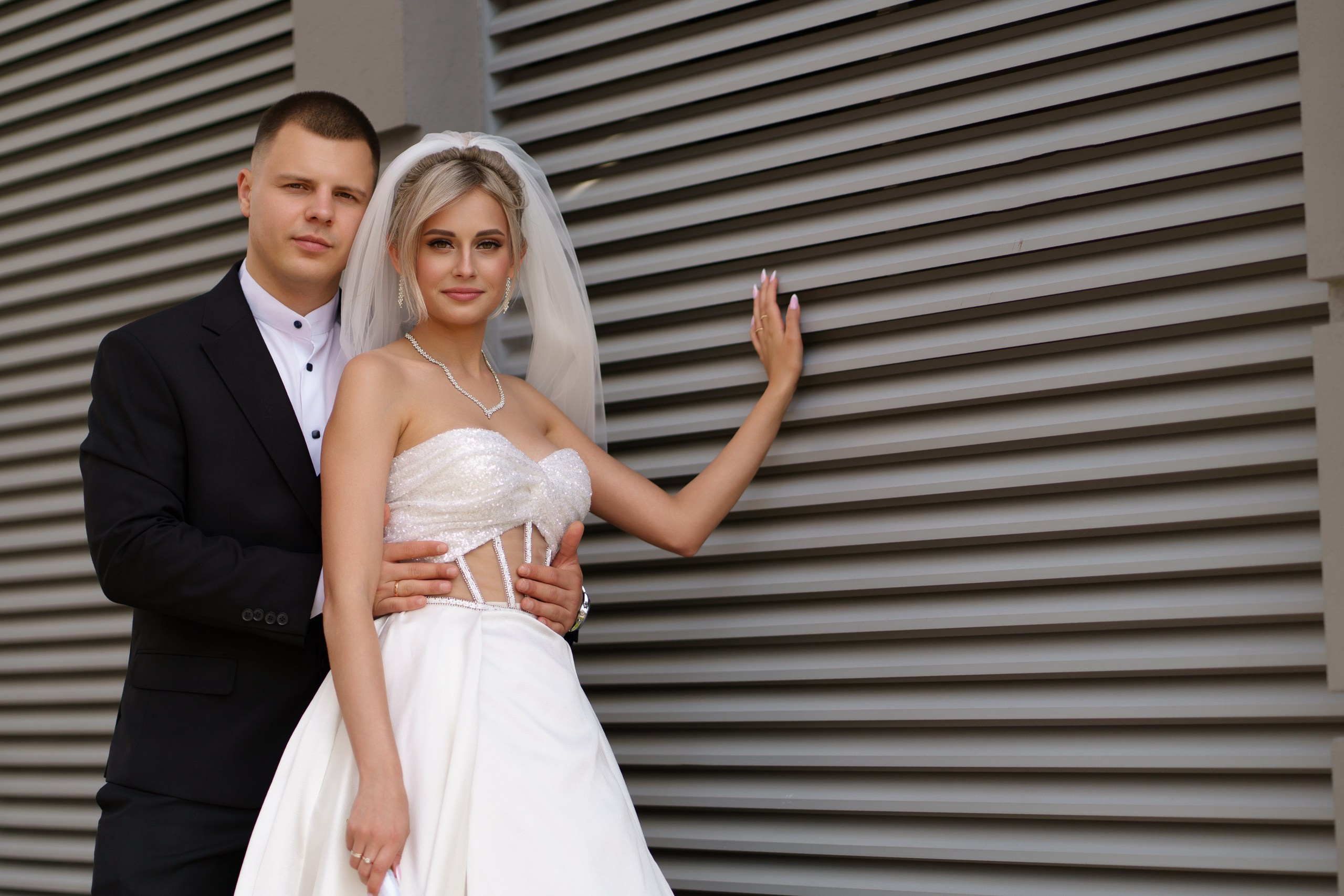 Свадебная пара на фоне современной стены с горизонтальными рифлёными панелями: жених в строгом чёрном костюме, невеста — в роскошном белом платье с корсетным верхом, фатой и жемчужным ожерельем | классический свадебный фотограф Екатеринбург