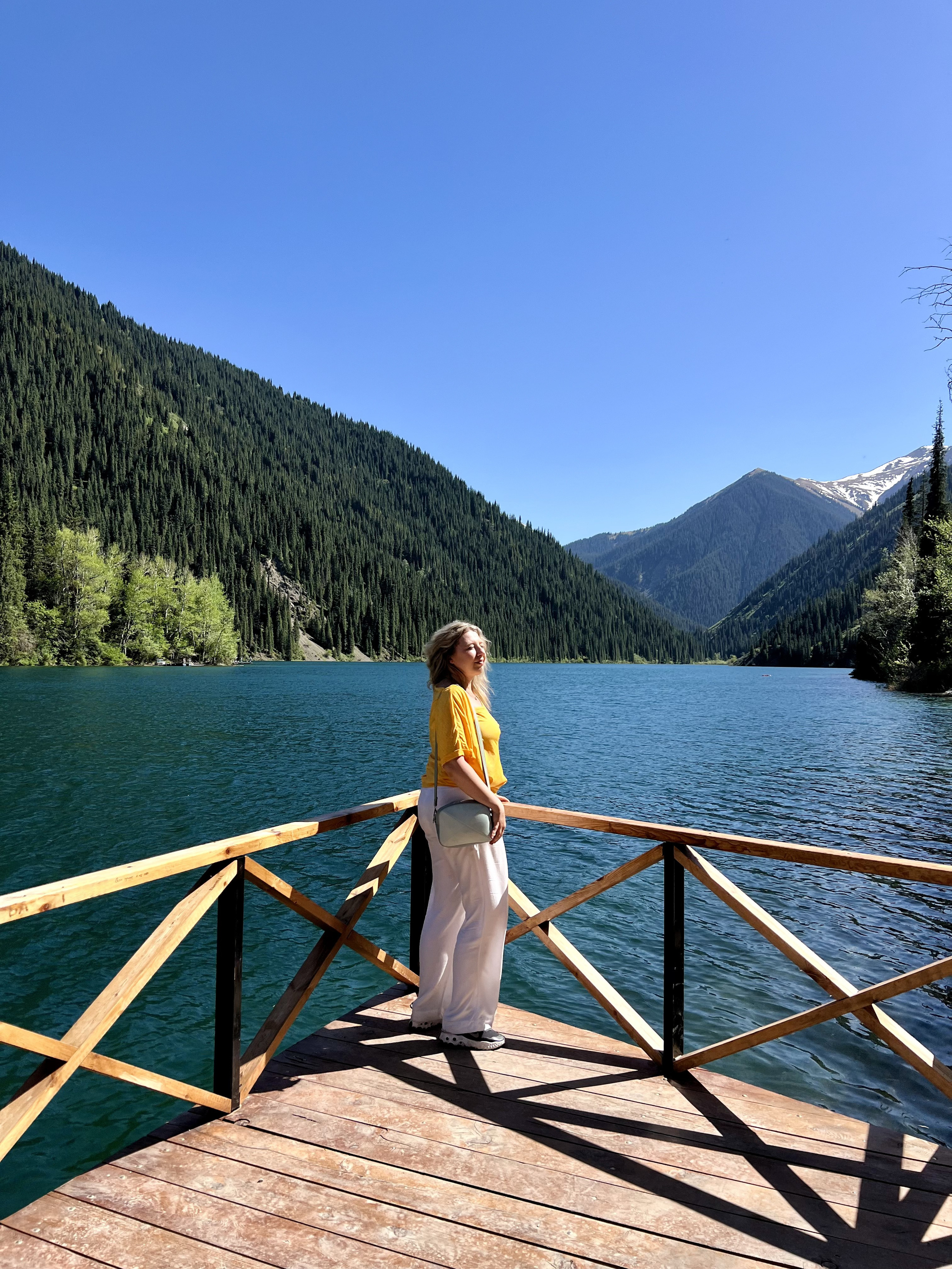 Женщина в ярко-жёлтой кофте и светлых брюках стоит на деревянной террасе с ограждением, выходящей на красивое горное Кольсайское озеро. Вода насыщенного синего цвета, а вокруг озера тянутся густо покрытые лесом горные склоны. На заднем плане видны величественные горы с заснеженными вершинами. Чистое голубое небо подчёркивает атмосферу солнечного, тёплого дня.