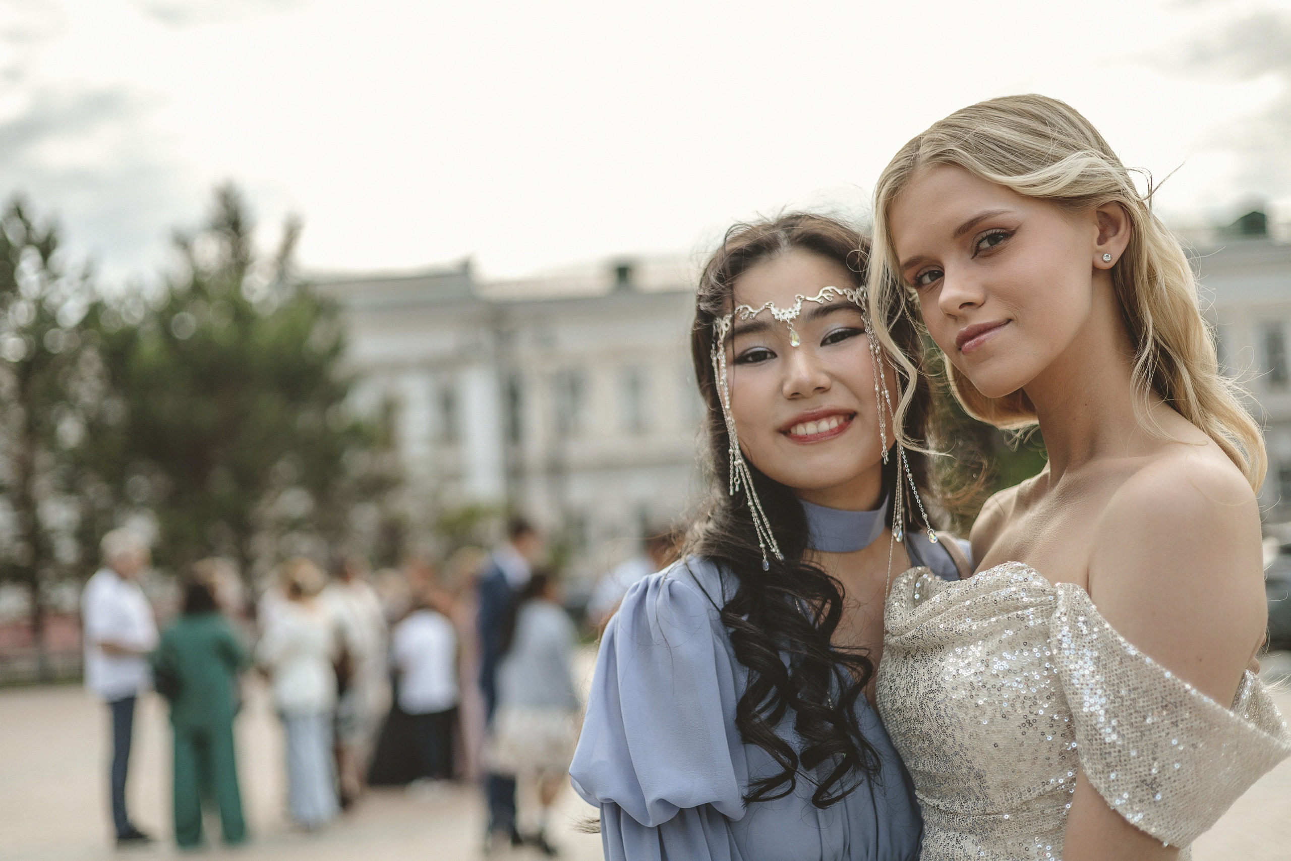 Две школьницы-выпускницы позируют на улице, улыбаясь. На одной из них блестящее платье с открытыми плечами, на другой - платье цвета лаванды с украшенным драгоценными камнями головным убором. На заднем плане люди и деревья.