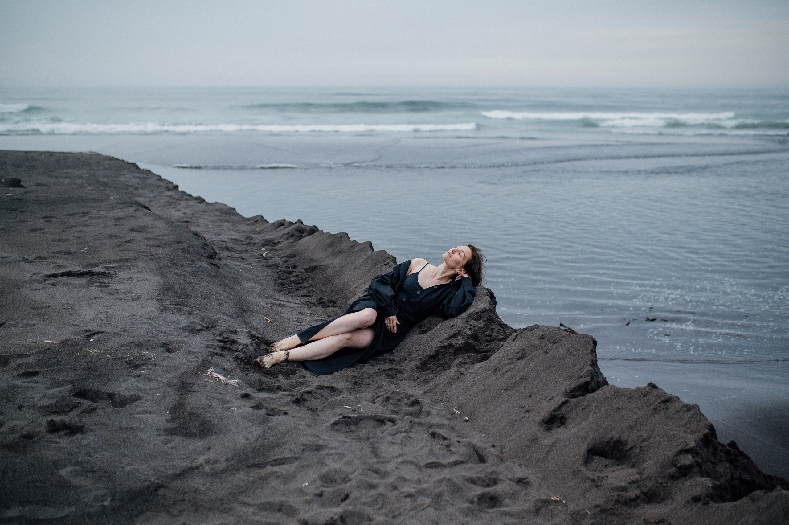 Девушка в тёмном платье полулежит на чёрном песке у моря, смотрит вверх. Атмосфера спокойная, немного меланхоличная.