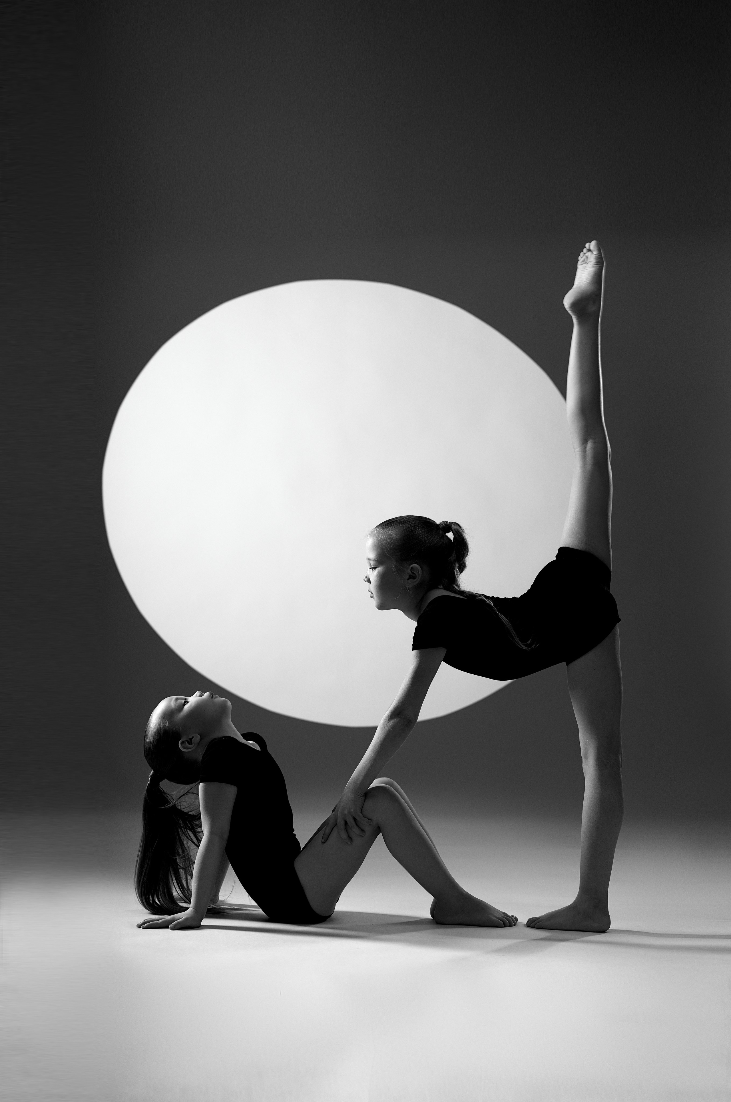 Художественный чб портрет двух гимнасток в студии на фоне светящегося диска. Эстетика линий и гибкости в жанре спорт-арт.