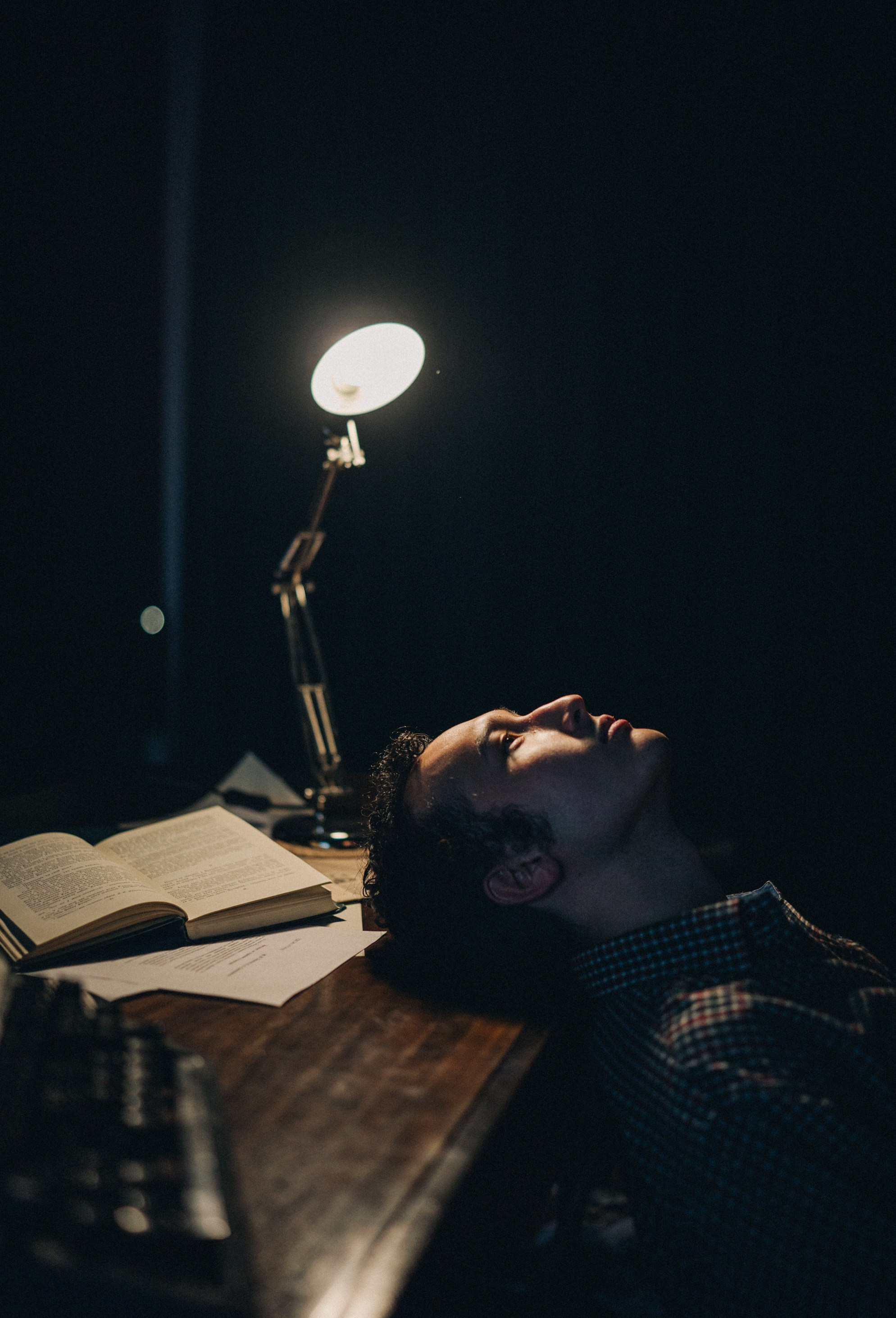 Мужчина в клетчатой рубашке сидит за деревянным столом в темной комнате, освещённый настольной лампой