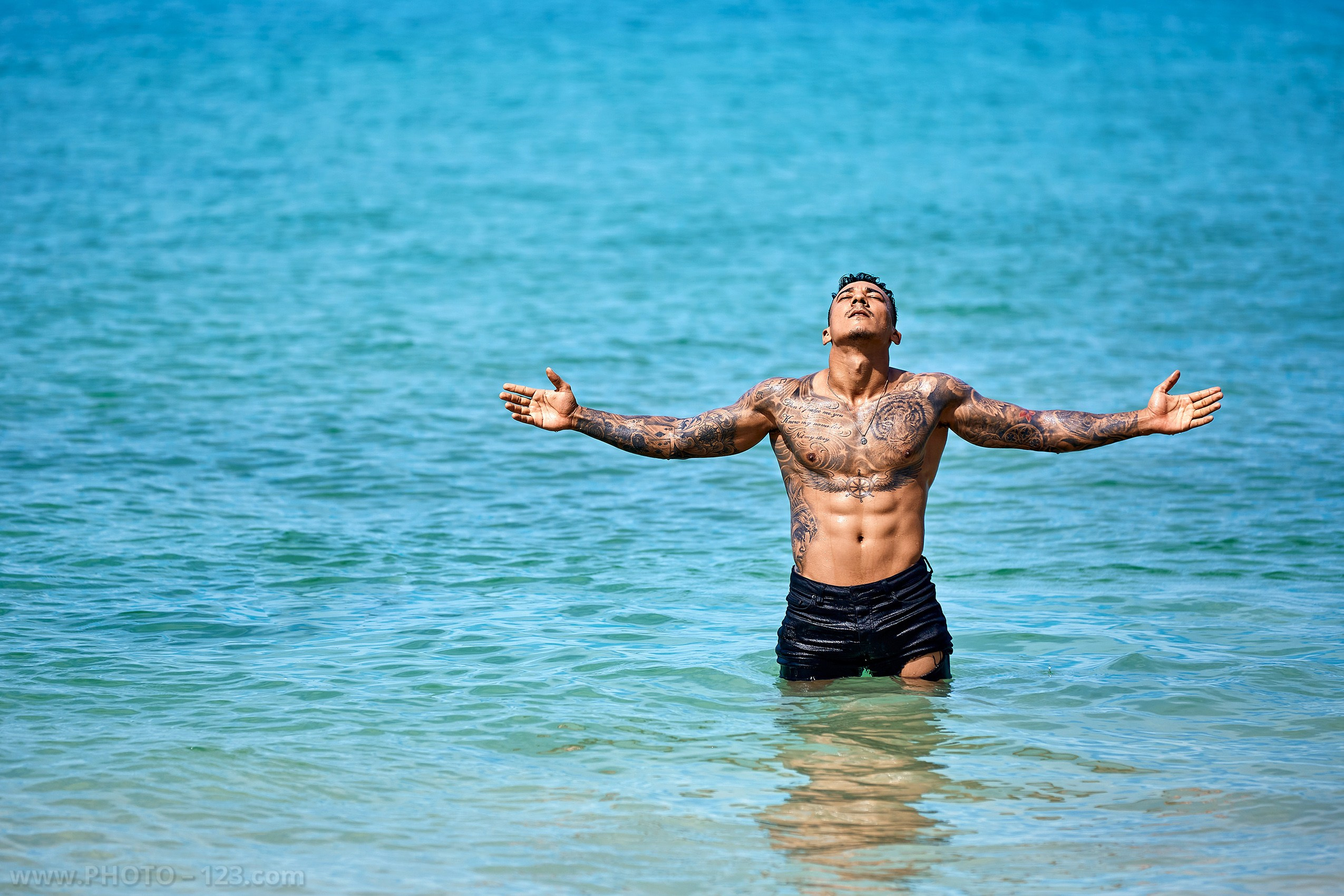 Muscular tattooed man standing waist-deep in the ocean with arms open on a Nha Trang beach