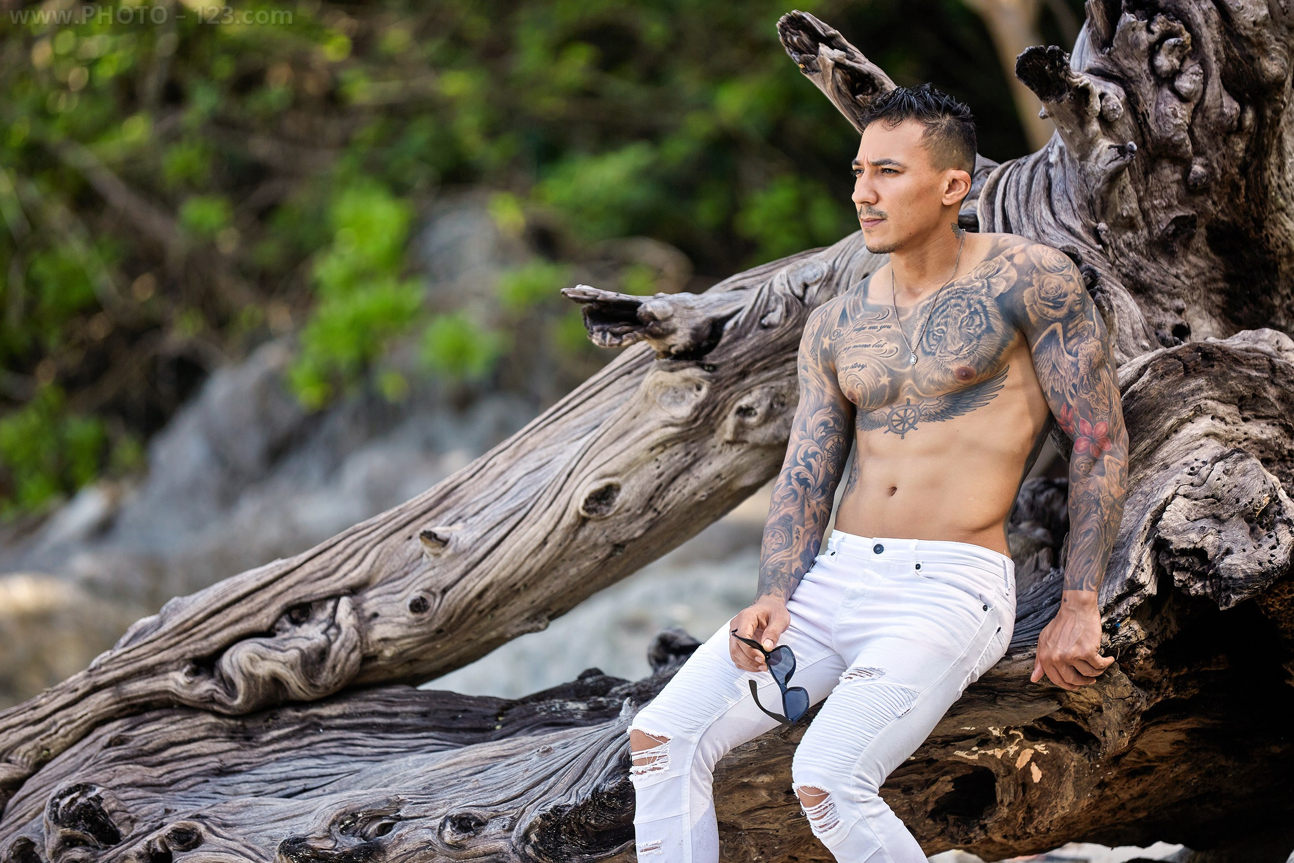 Shirtless athletic model holding sunglasses on a tree trunk during a Nha Trang beach photoshoot