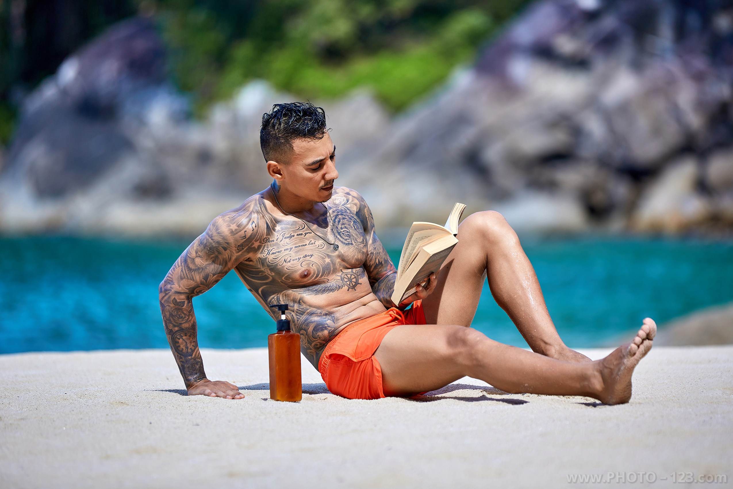 Tattooed muscular man reclining on white sand with a book during a beach photoshoot in Nha Trang