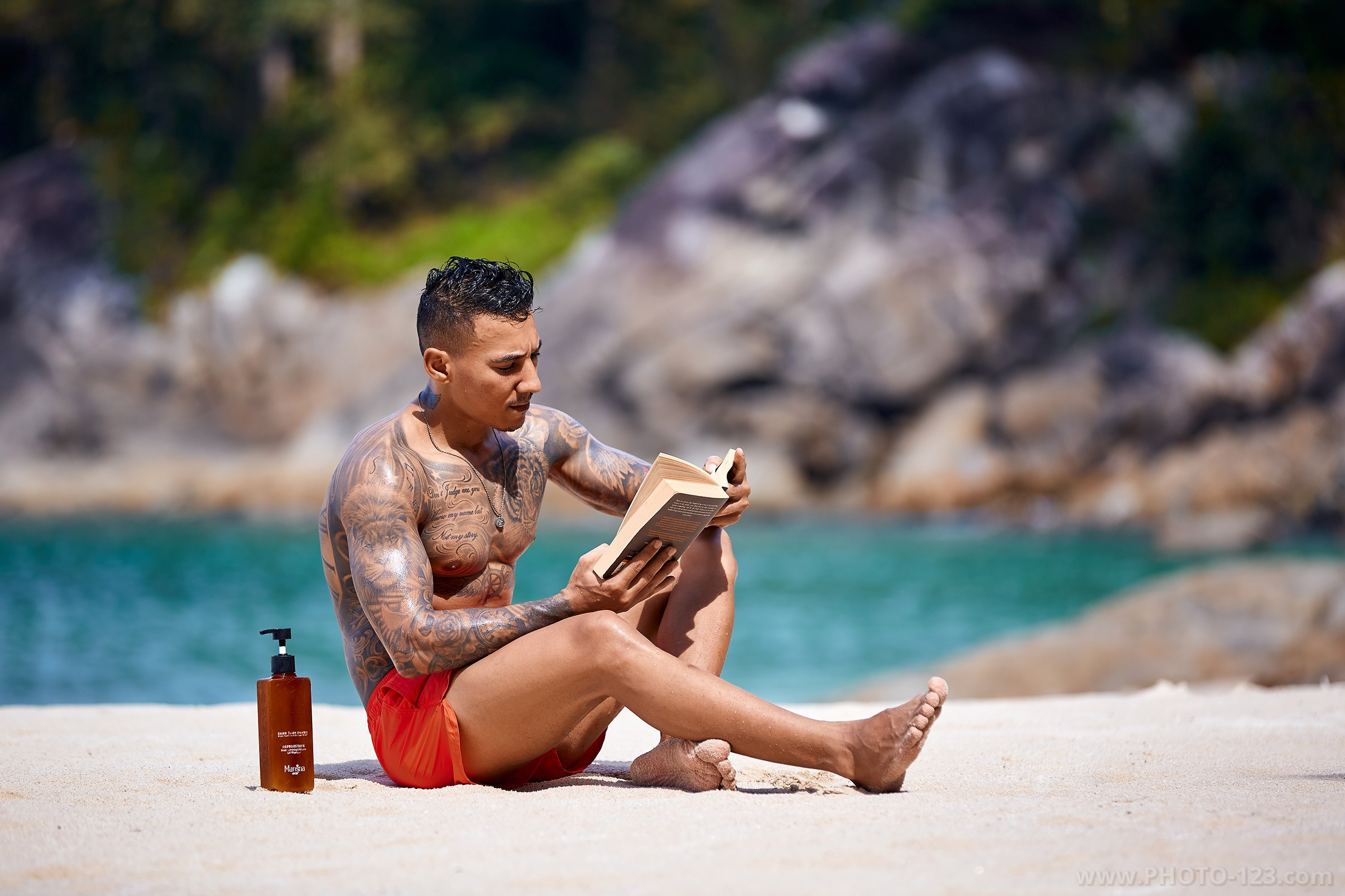 Shirtless tattooed male model sitting on white beach sand reading a book against turquoise sea in Nha Trang captured by professional photographer