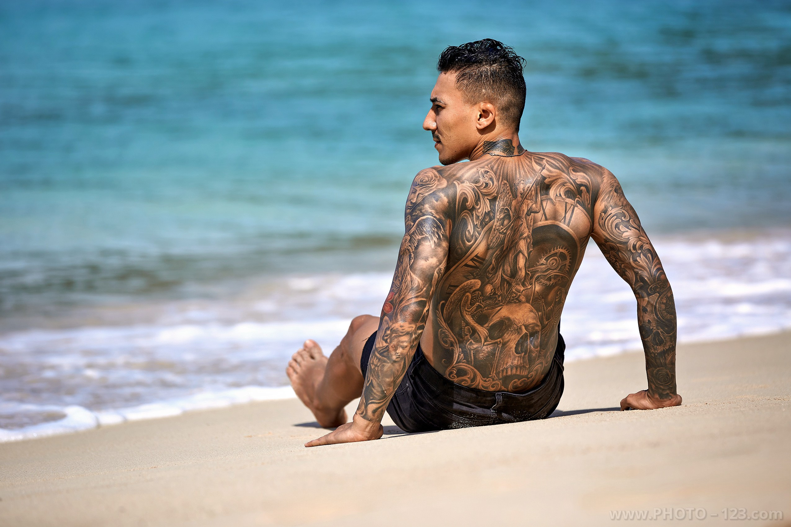 Back view of a tattooed muscular man sitting on the beach shore by Nha Trang photographer