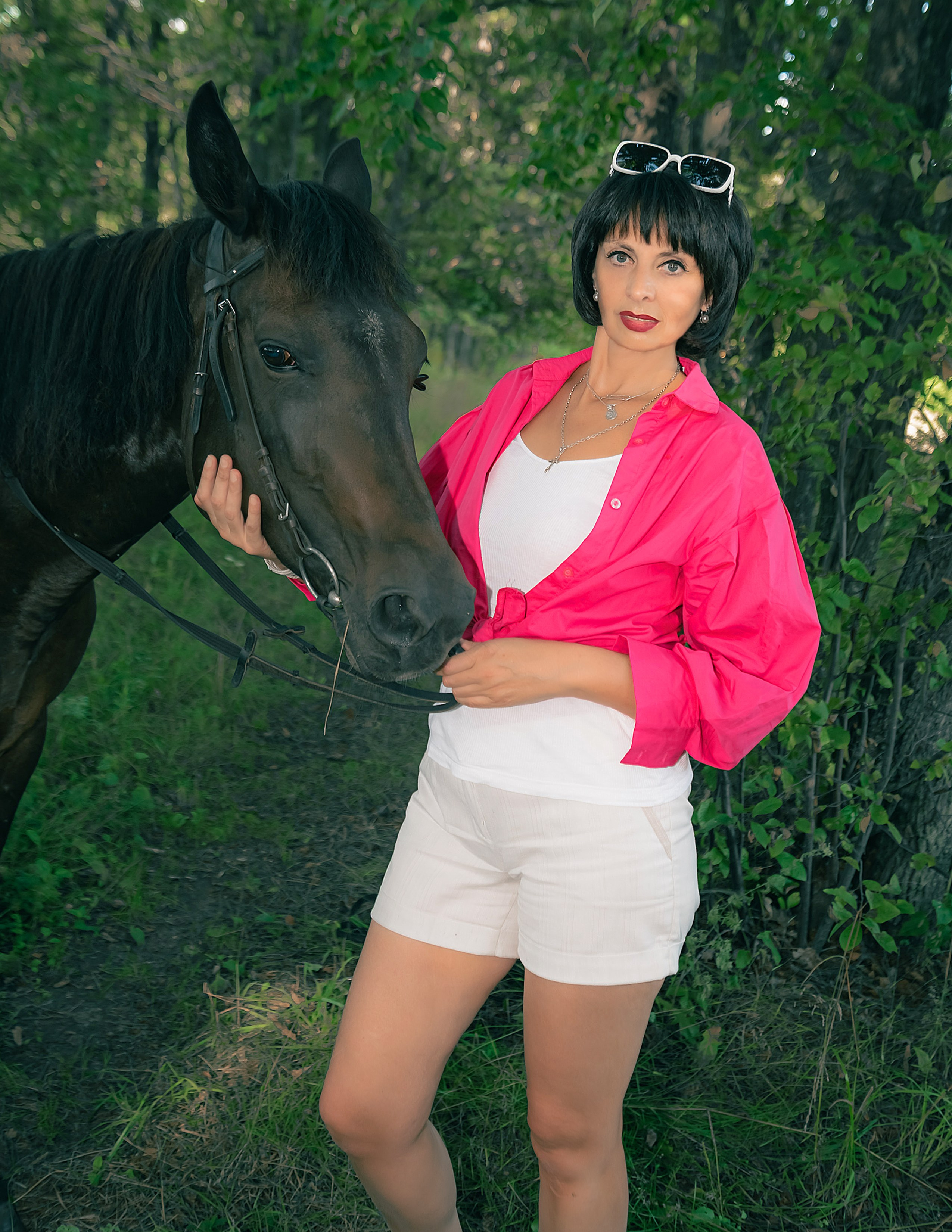 На фото женщина в рубашке стоит с лошадью на опушке леса.