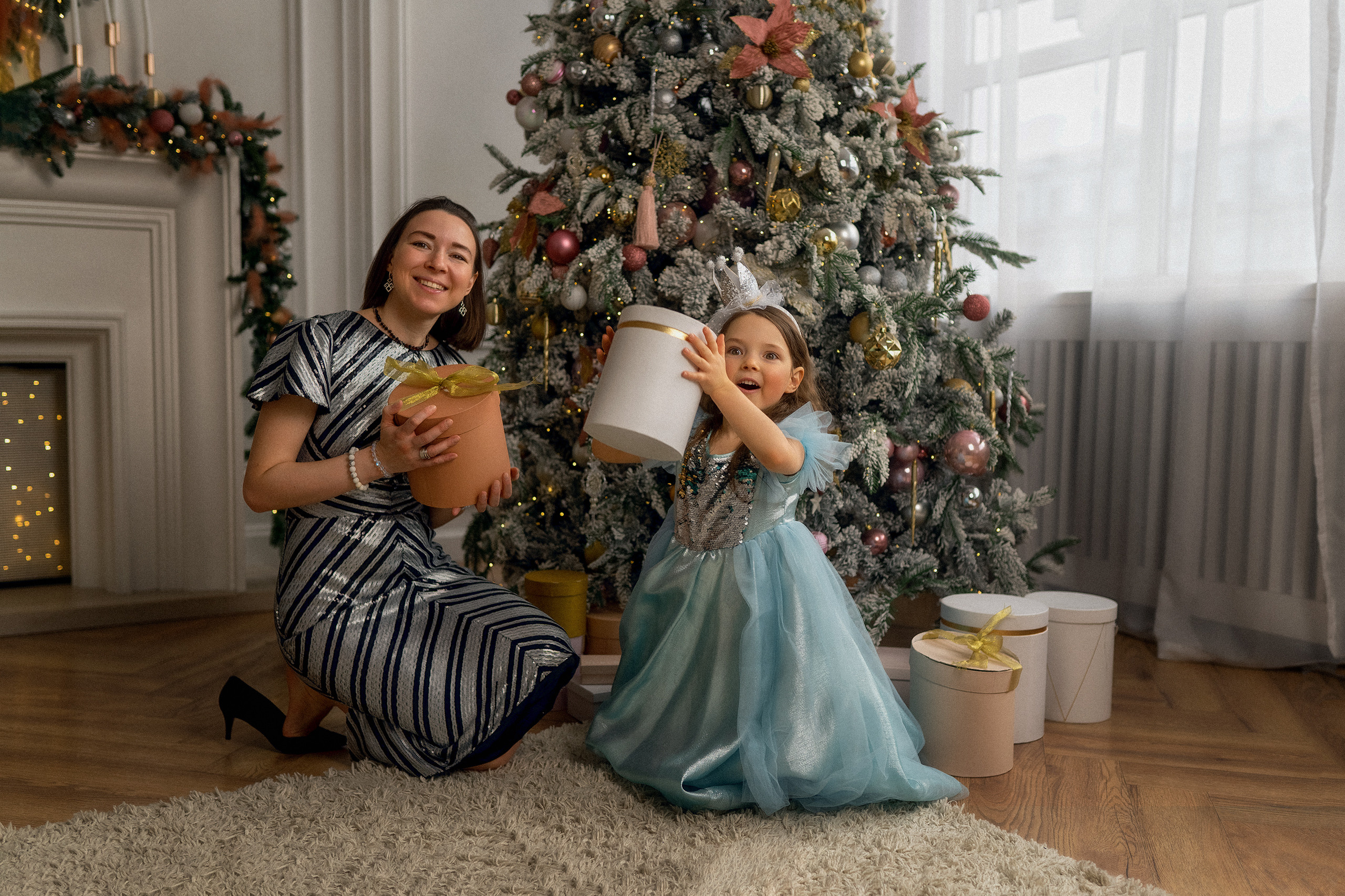 Маленькая девочка в платье принцессы с короной на головерядом с мамой держат в руках подарки на фоне камина новогодней елки и подарочных коробок